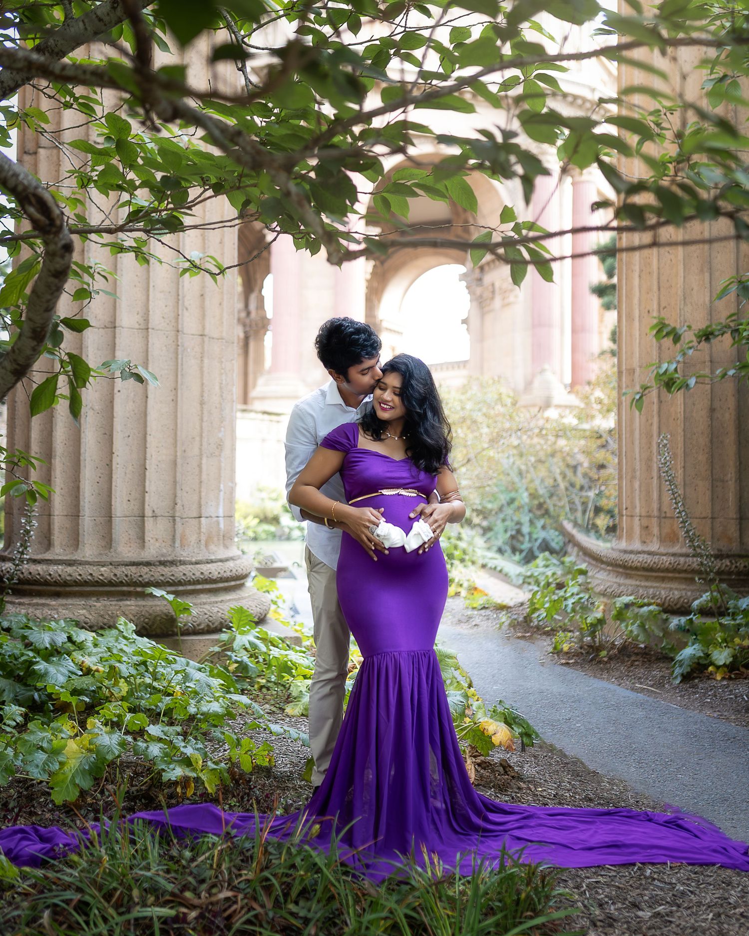 A couple in formal attire embrace on a garden path with stone columns and a flowing purple dress train.