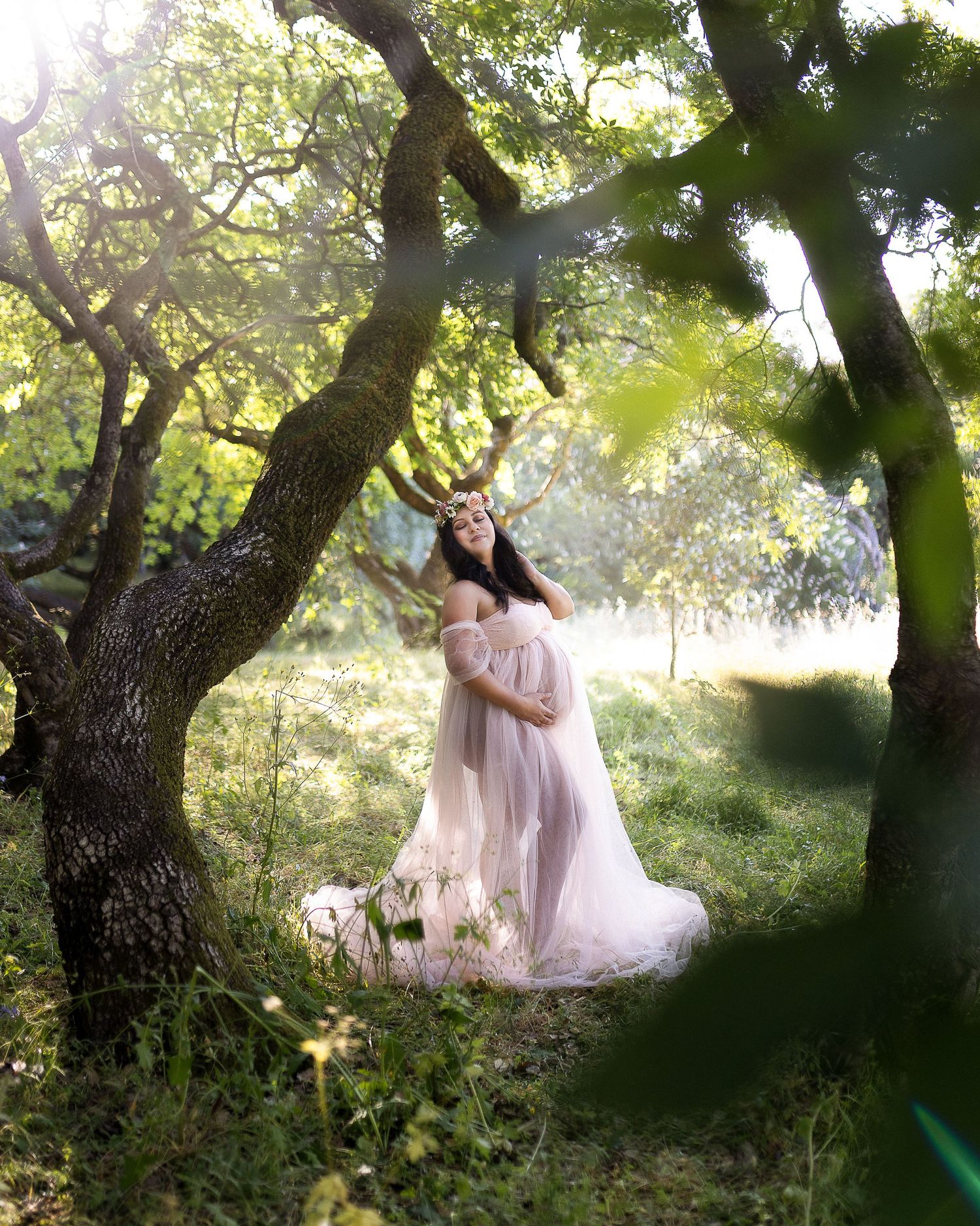 Dreamy maternity photo in a sunlit forest with flowing pink dress and natural tree framing.