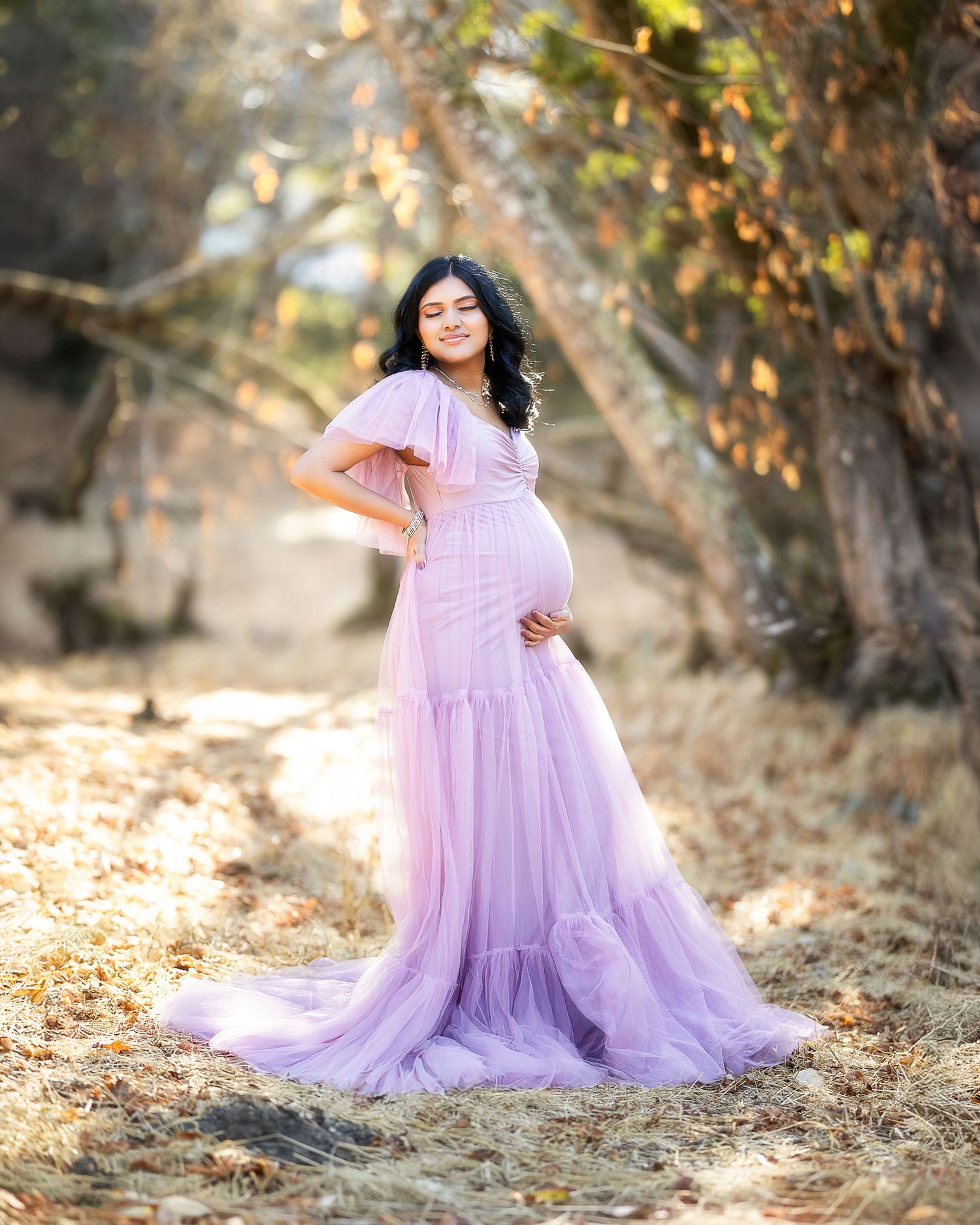 A flowing lavender tulle maternity gown captures an ethereal moment in a sunlit autumn setting with fallen leaves.