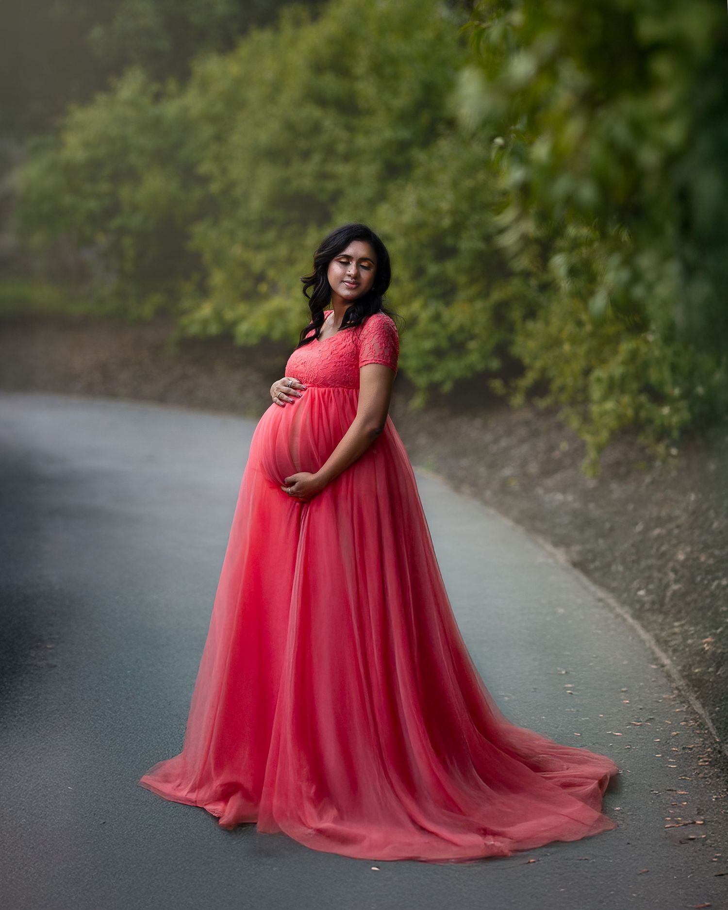 A stunning coral maternity dress billows gracefully in a natural outdoor setting with green foliage.