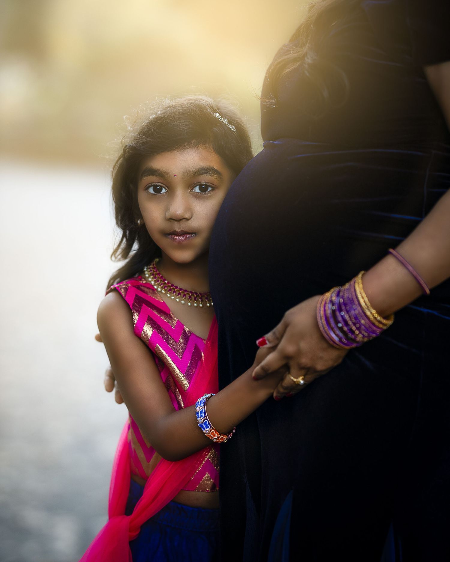 A young girl in a bright pink dress stands next to someone wearing dark clothing in soft lighting.