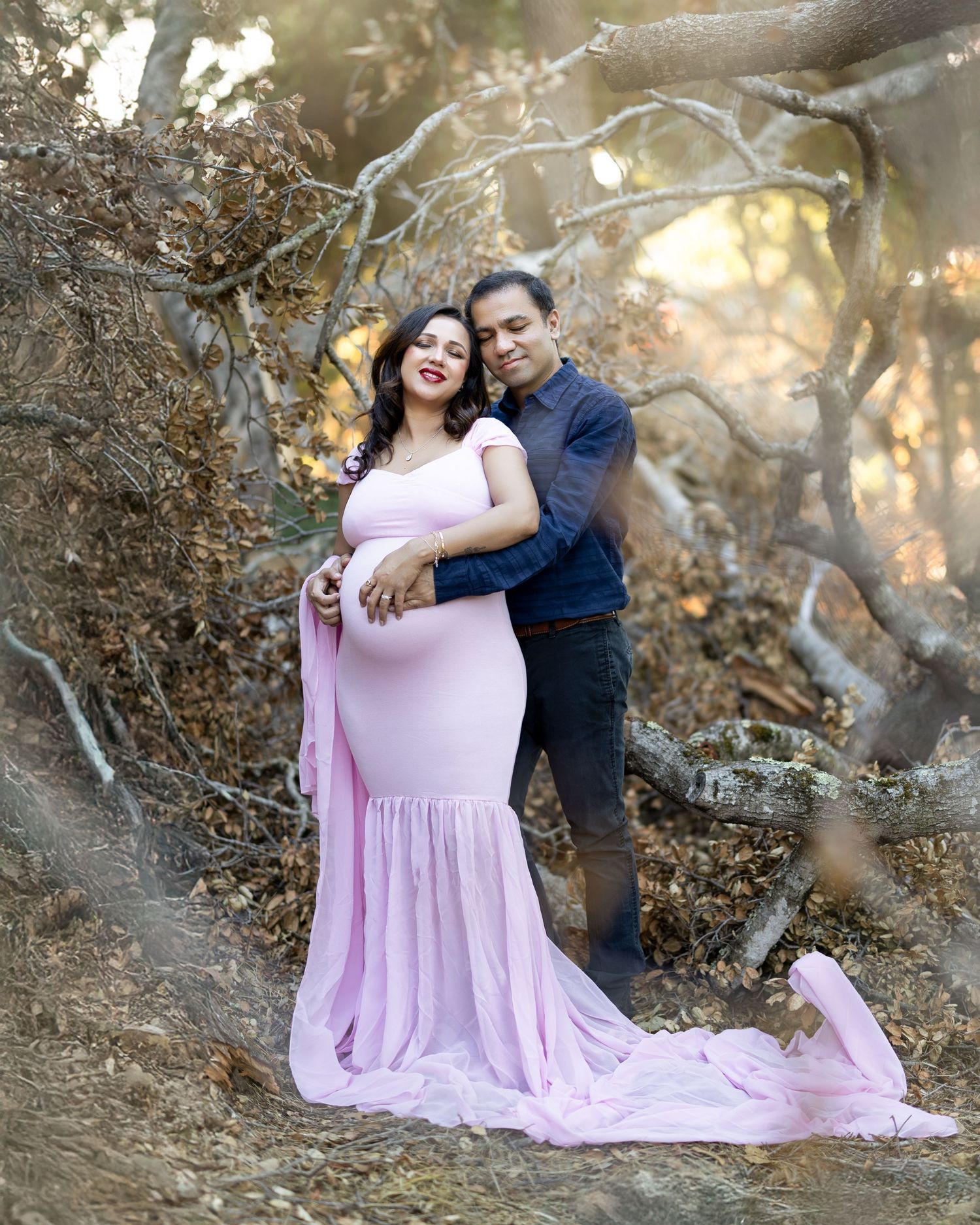 A couple in a romantic outdoor photoshoot with a pink flowing dress against a dreamy forest backdrop during golden hour.