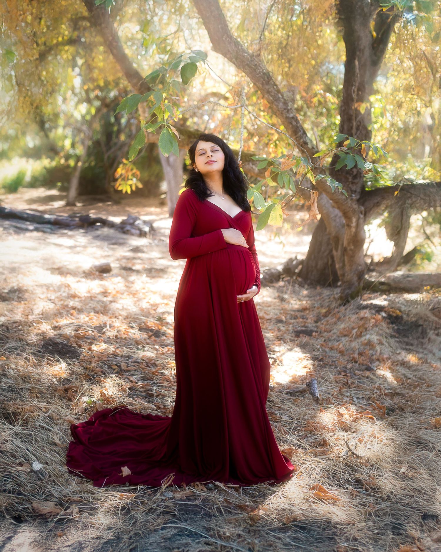 A figure in a flowing burgundy dress poses gracefully among autumn trees in a dreamy outdoor maternity photoshoot.