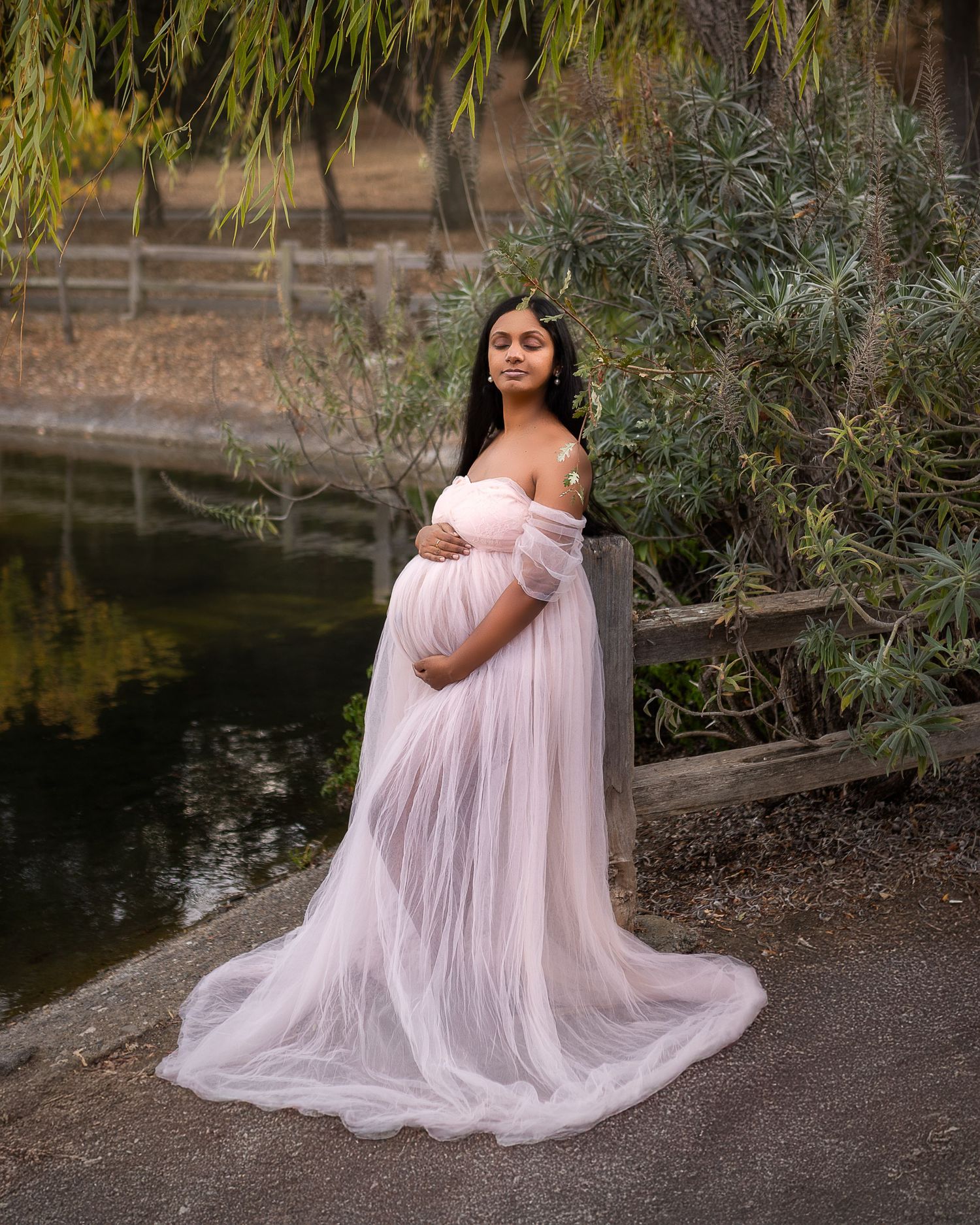 Soft pink tulle dress creates ethereal effect in outdoor maternity photos beside a tranquil pond.