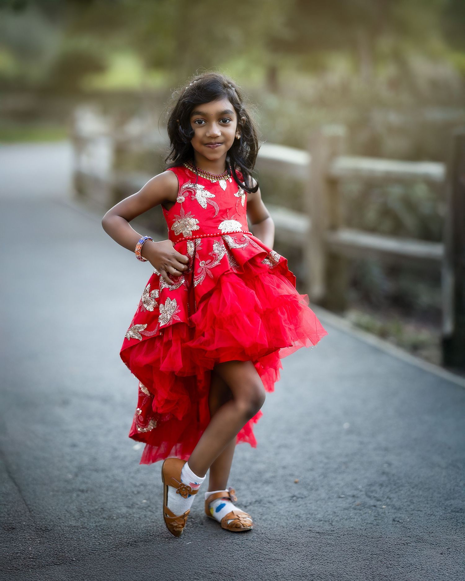 A child in a bright red floral high-low party dress with tulle layers poses on a path outdoors.