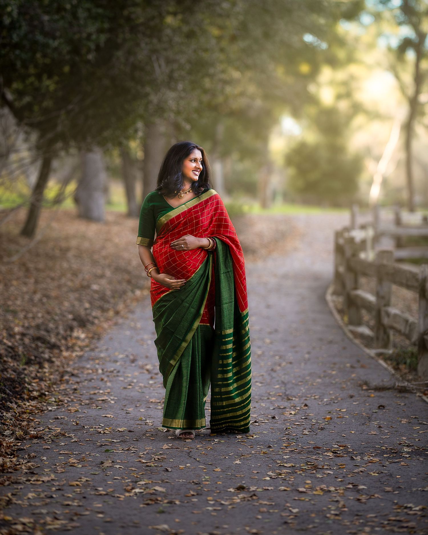 A flowing red and green sari dress billows gracefully along an autumn path lined with fallen leaves and wooden fencing.