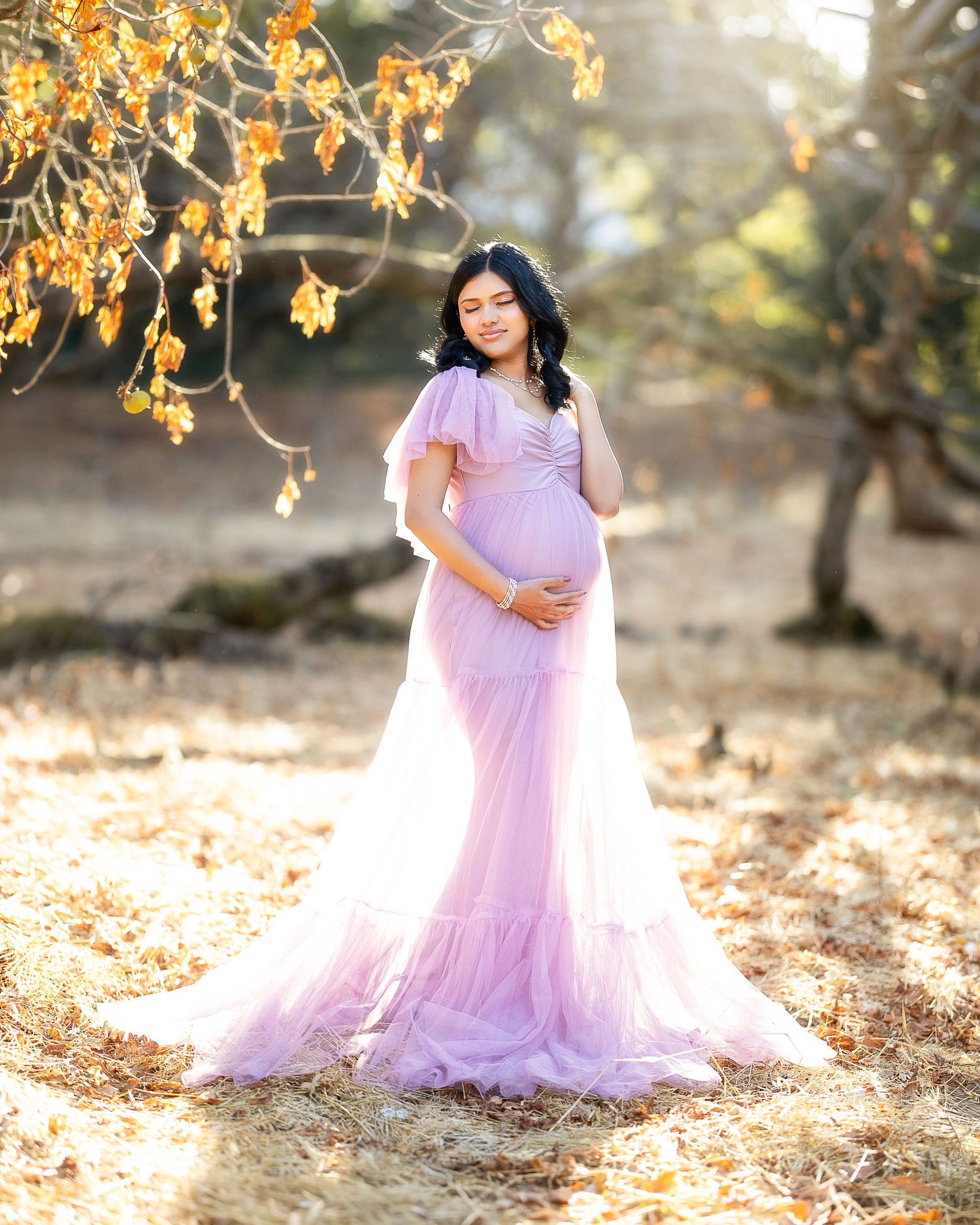 A maternity photo of a figure in a flowing lavender gown standing beneath autumn leaves in soft golden light.