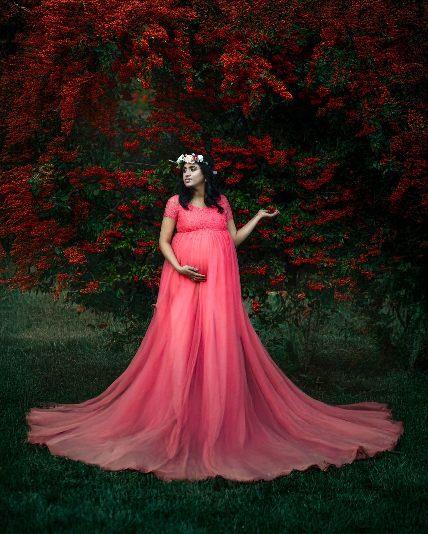 A figure in a flowing coral pink dress poses gracefully against a backdrop of dark red autumn foliage in a forest setting.