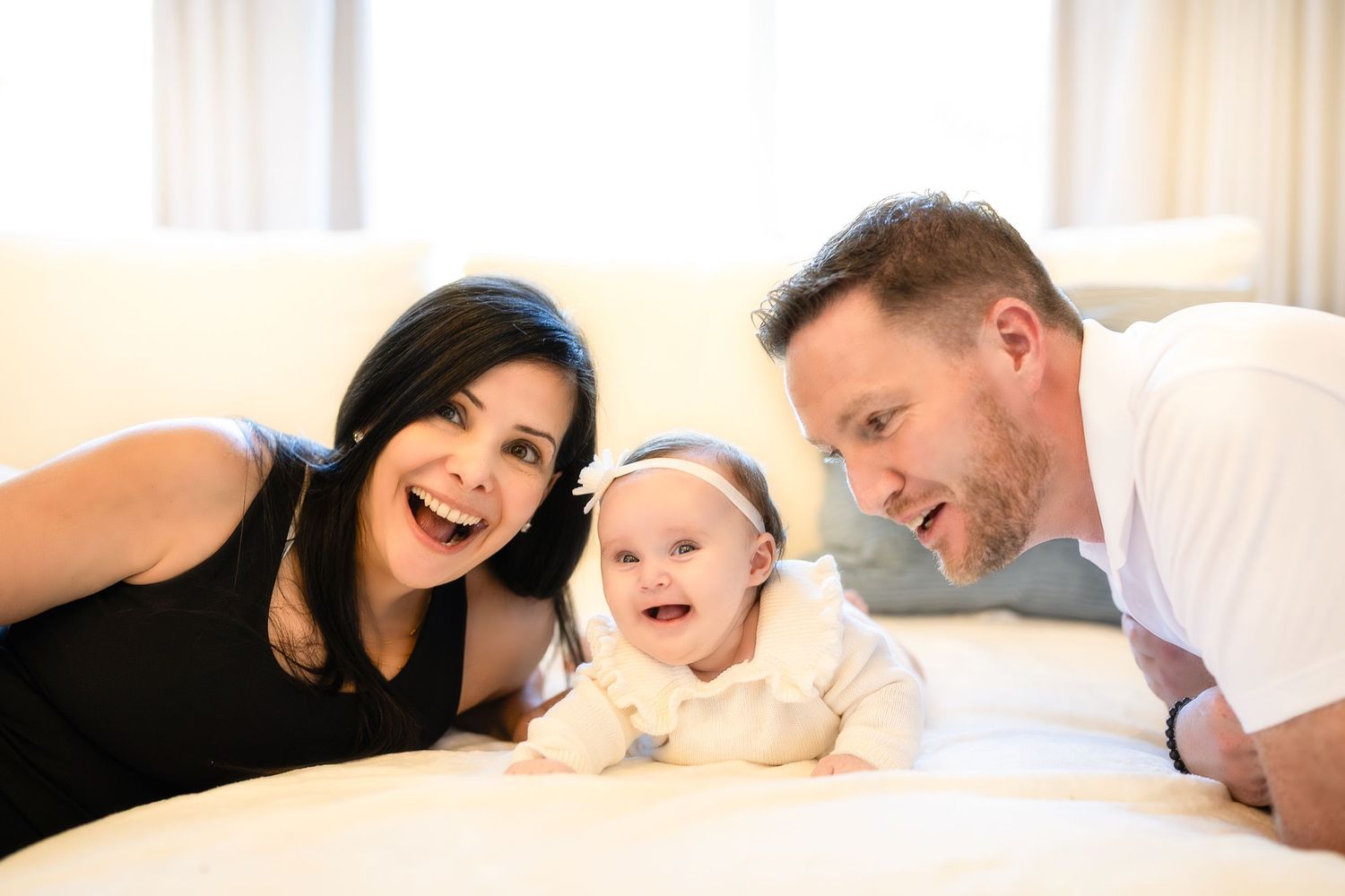 Happy family laughing together while laying on white bedding in a bright room.