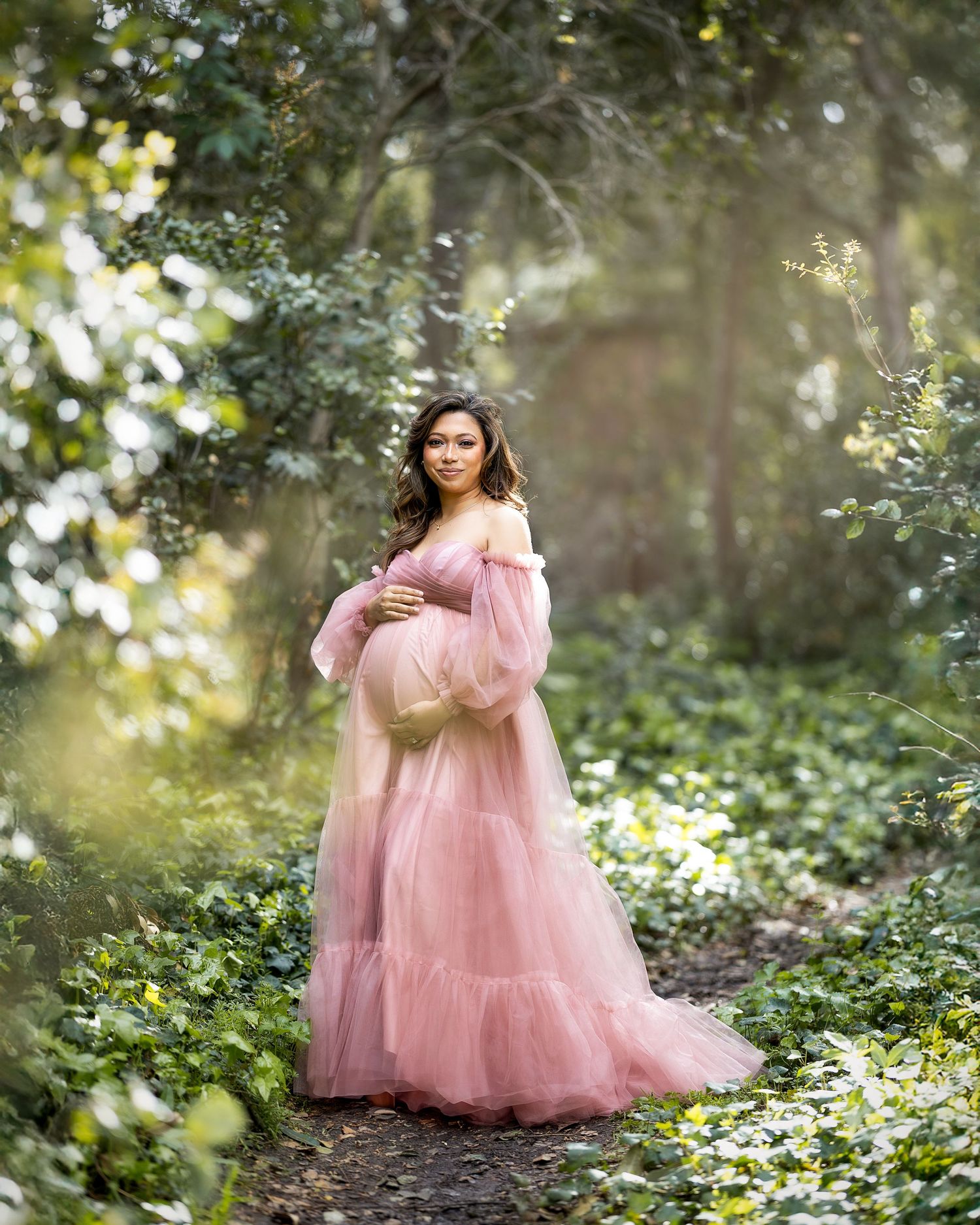 A dreamy maternity photo shows a woman in a flowing pink tulle gown standing in a sunlit forest surrounded by flowers.