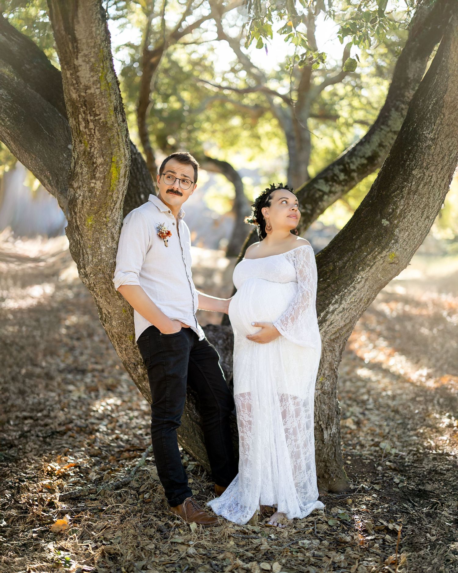A couple poses playfully against a forked tree trunk in a natural outdoor setting, with the person in a white lace maternity dress.
