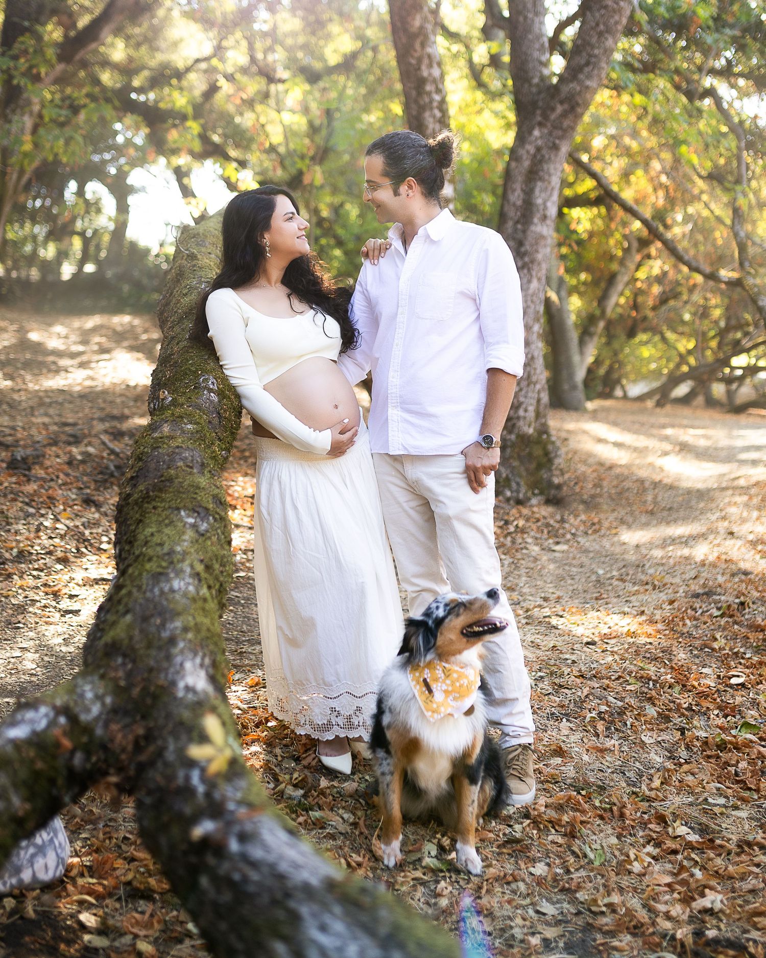 A couple in white outfits share a tender moment in a wooded setting with their Australian Shepherd dog beside them.