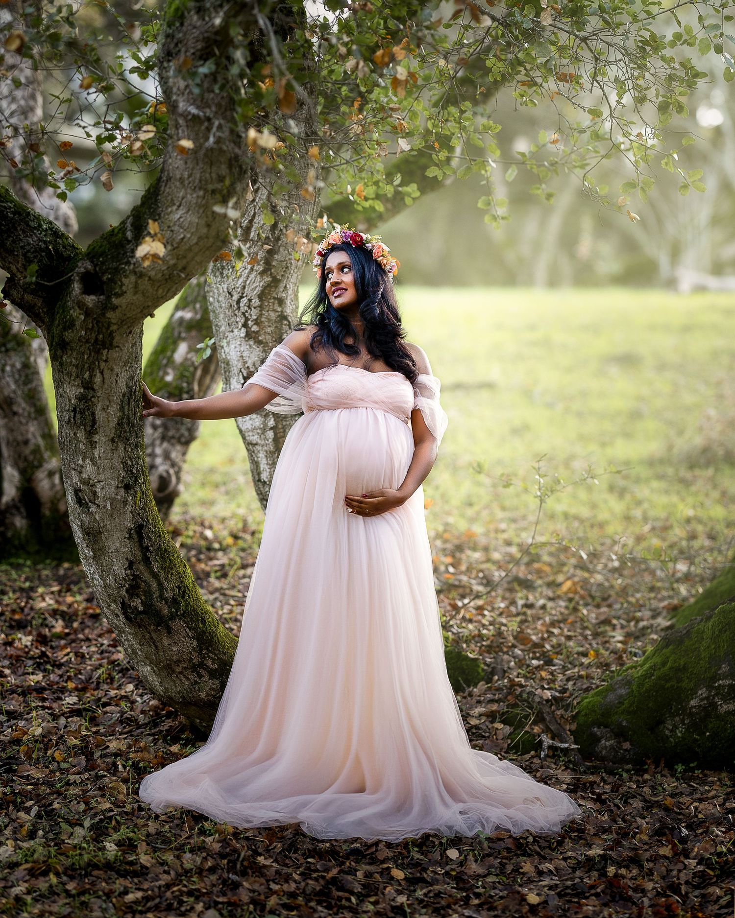 A maternity photoshoot in a dreamy outdoor setting with a flowing pink tulle gown under autumn trees.