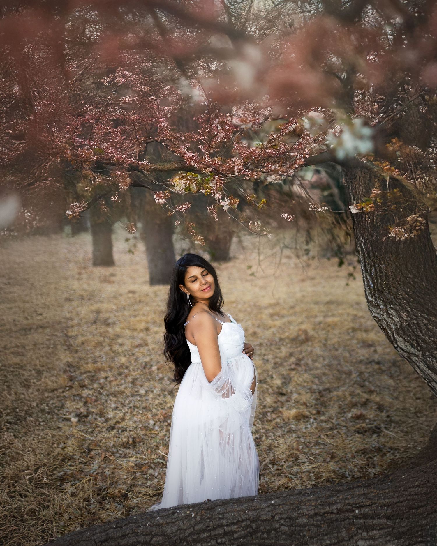 A woman in a white dress poses under blooming pink cherry blossom trees during a dreamy outdoor photo session.