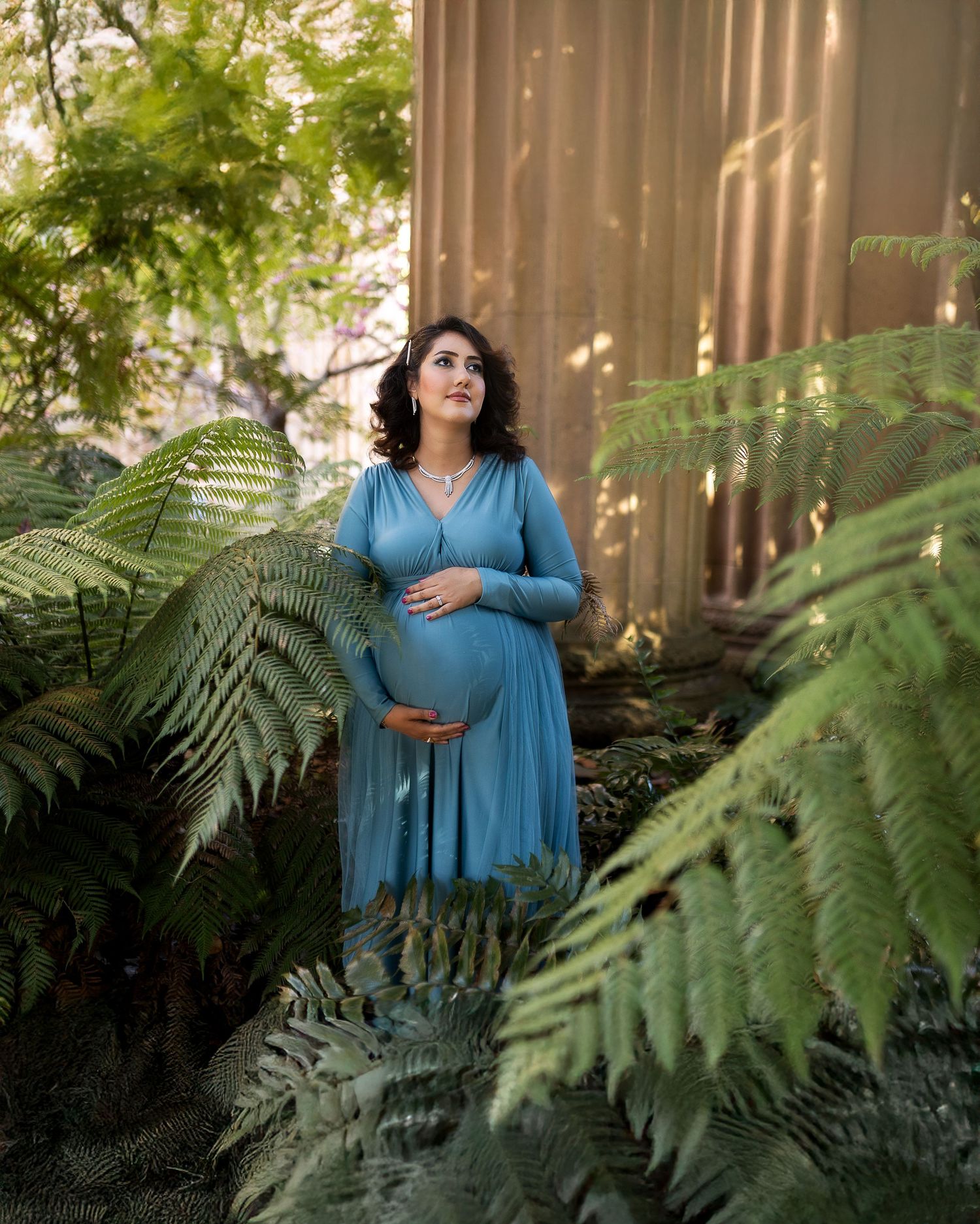 A figure in a blue dress poses gracefully among lush green ferns in dappled sunlight.