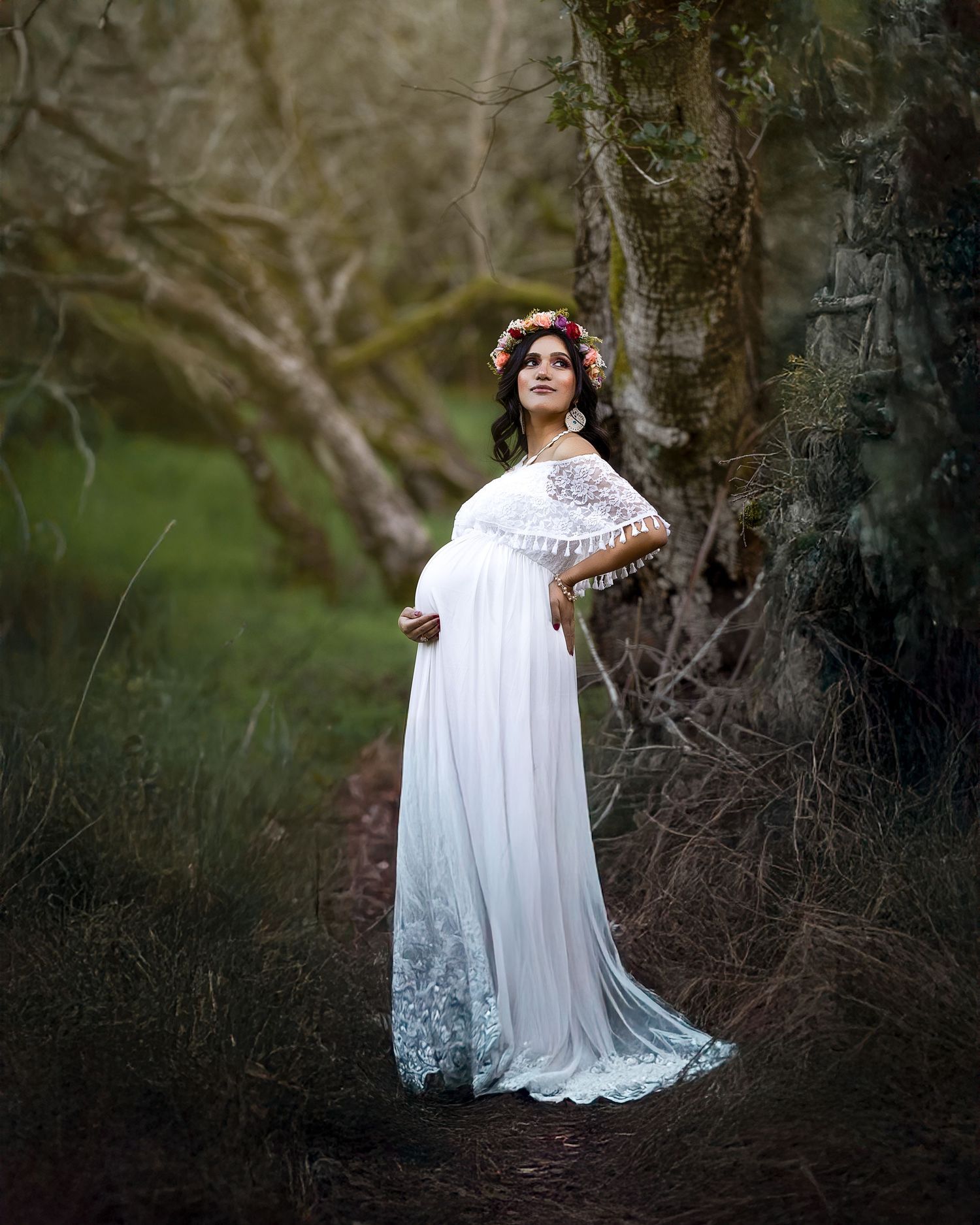 A person in a flowing white lace maternity gown poses elegantly in a wooded outdoor setting at dusk.