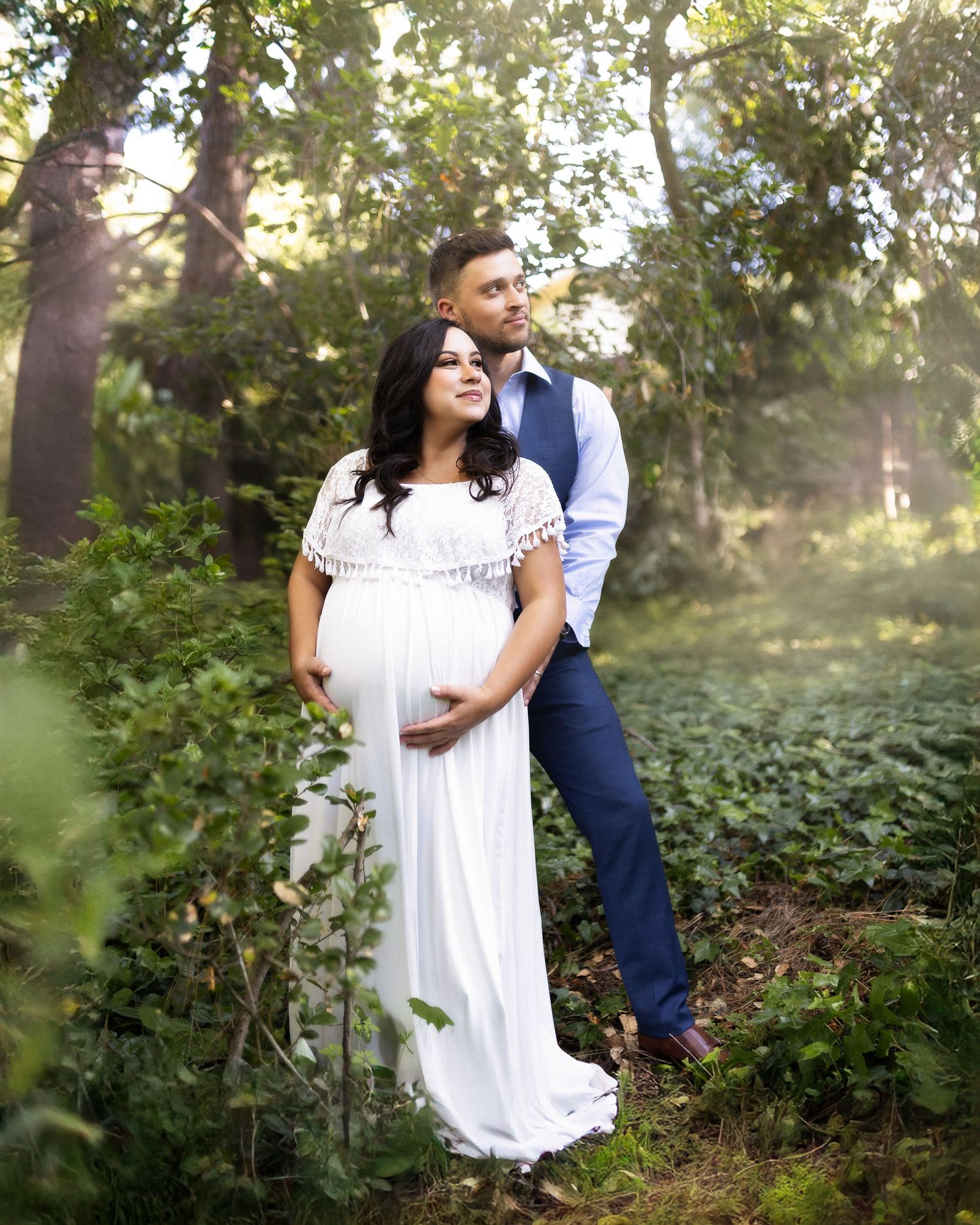 A couple in a romantic outdoor setting with a white dress and navy suit share tender moments in a wooded area.