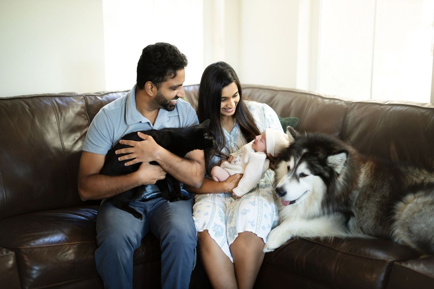 A couple sits on a brown leather couch with their Husky dog and black cat, sharing a tender moment at home.