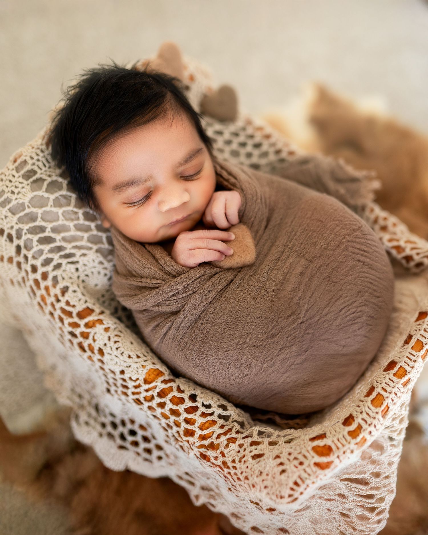 Sleeping newborn wrapped in brown swaddle resting on delicate white lace blanket in warm natural lighting.