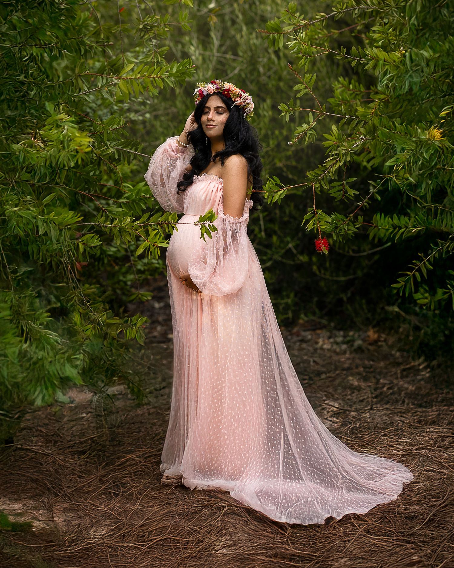 A figure in a flowing pink tulle dress and floral crown poses gracefully in a wooded outdoor setting.