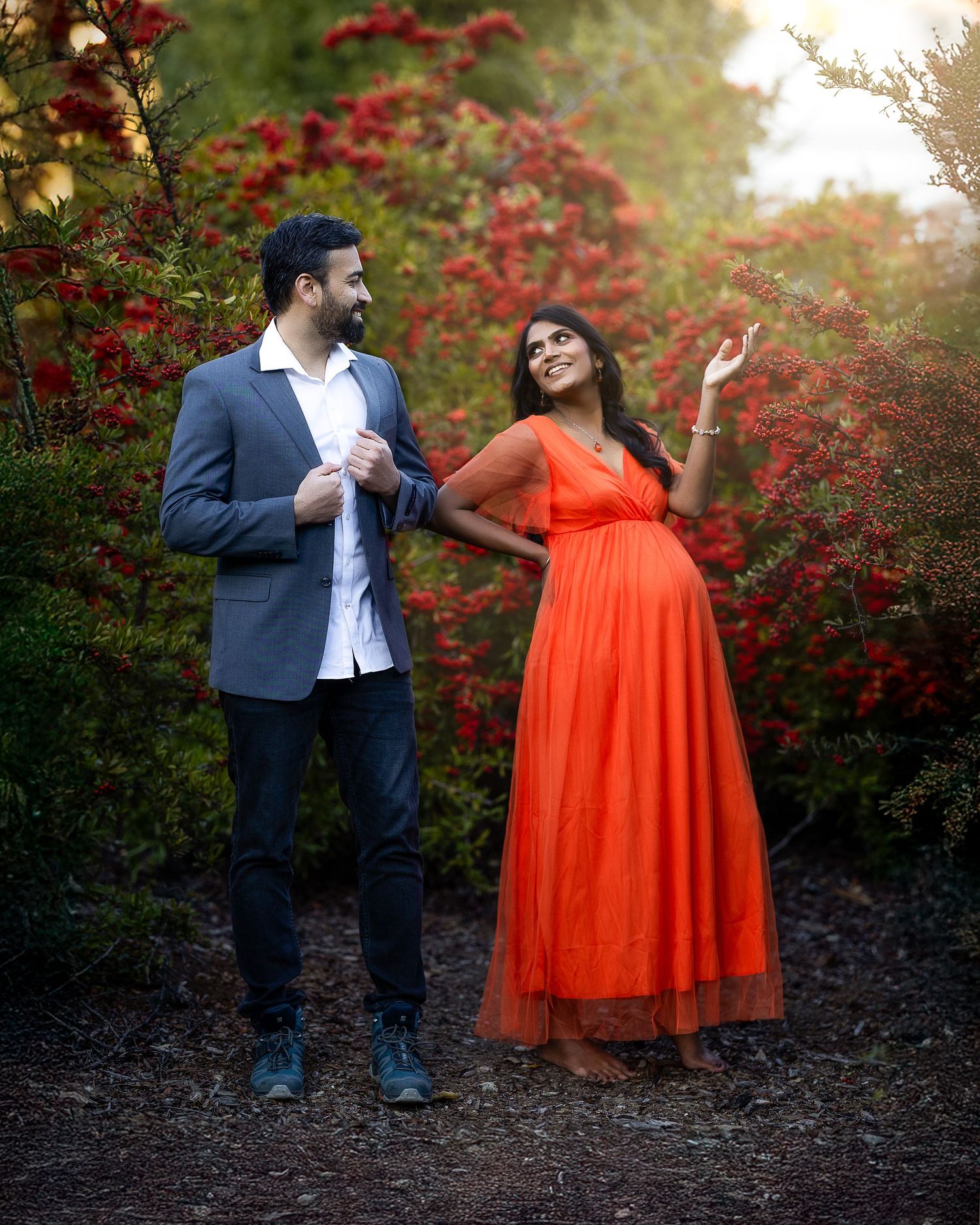A couple poses playfully against vibrant red autumn foliage, with her wearing a flowing orange gown.