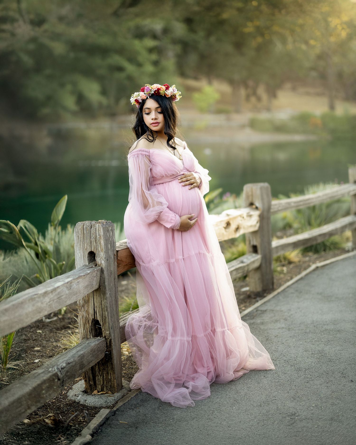 A flowing pink tulle maternity gown creates an ethereal scene along a peaceful lakeside path.