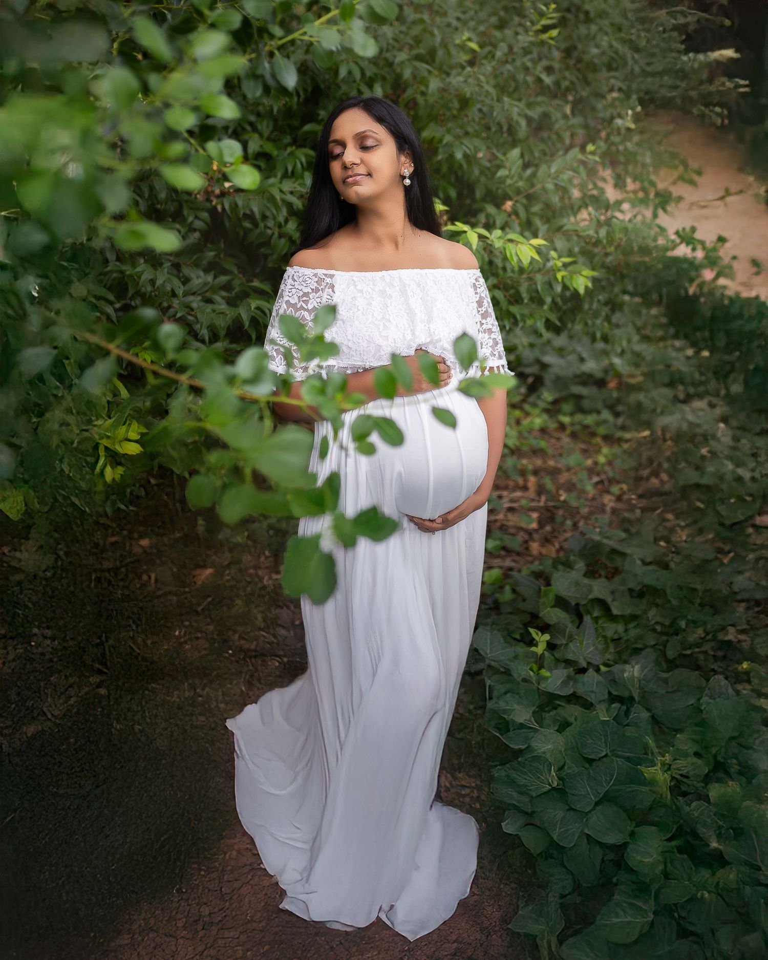 A person in a flowing white off-shoulder dress poses gracefully in a lush garden setting surrounded by green foliage.