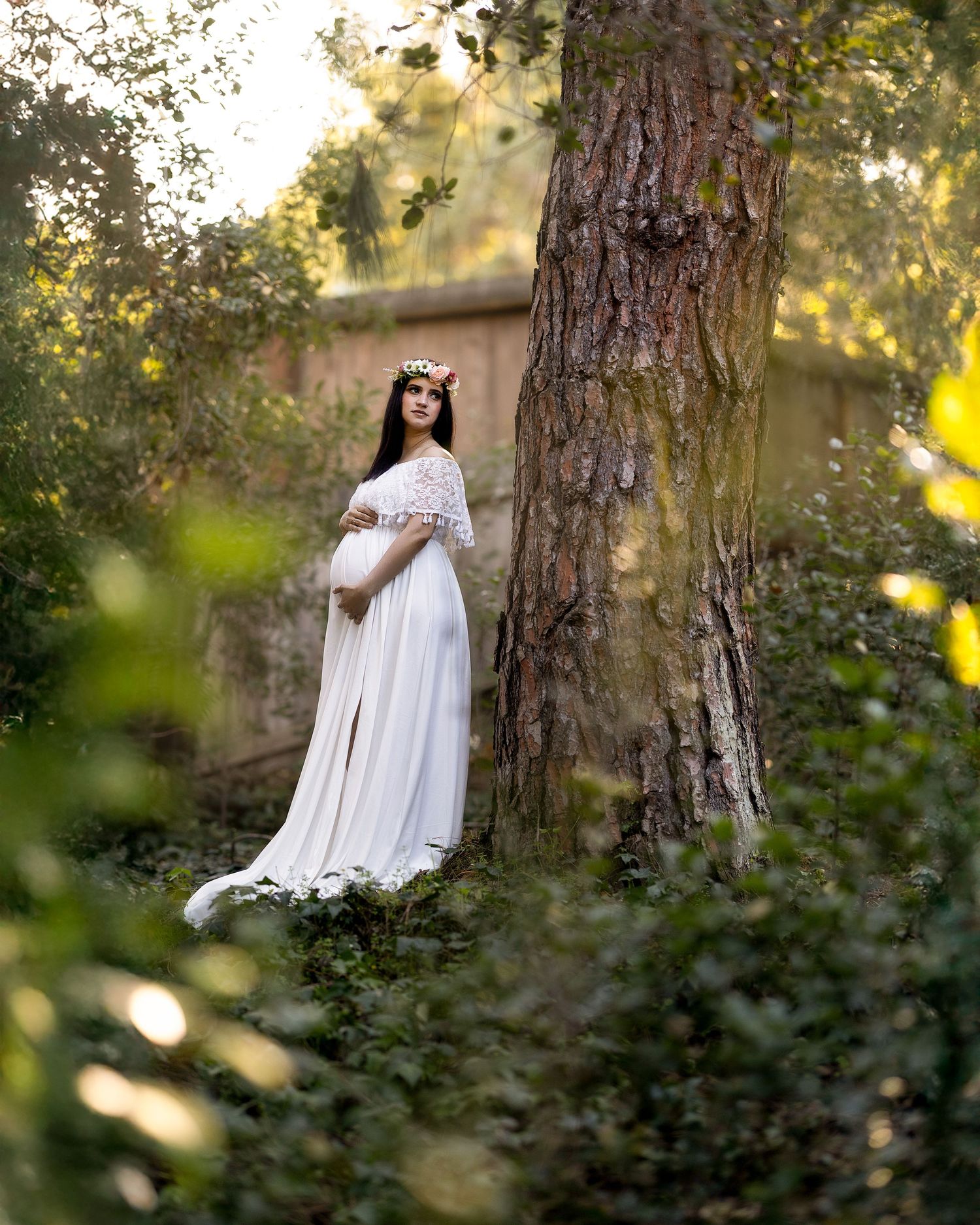 A dreamy maternity portrait in a white flowing dress standing beside a large tree trunk in dappled forest sunlight.