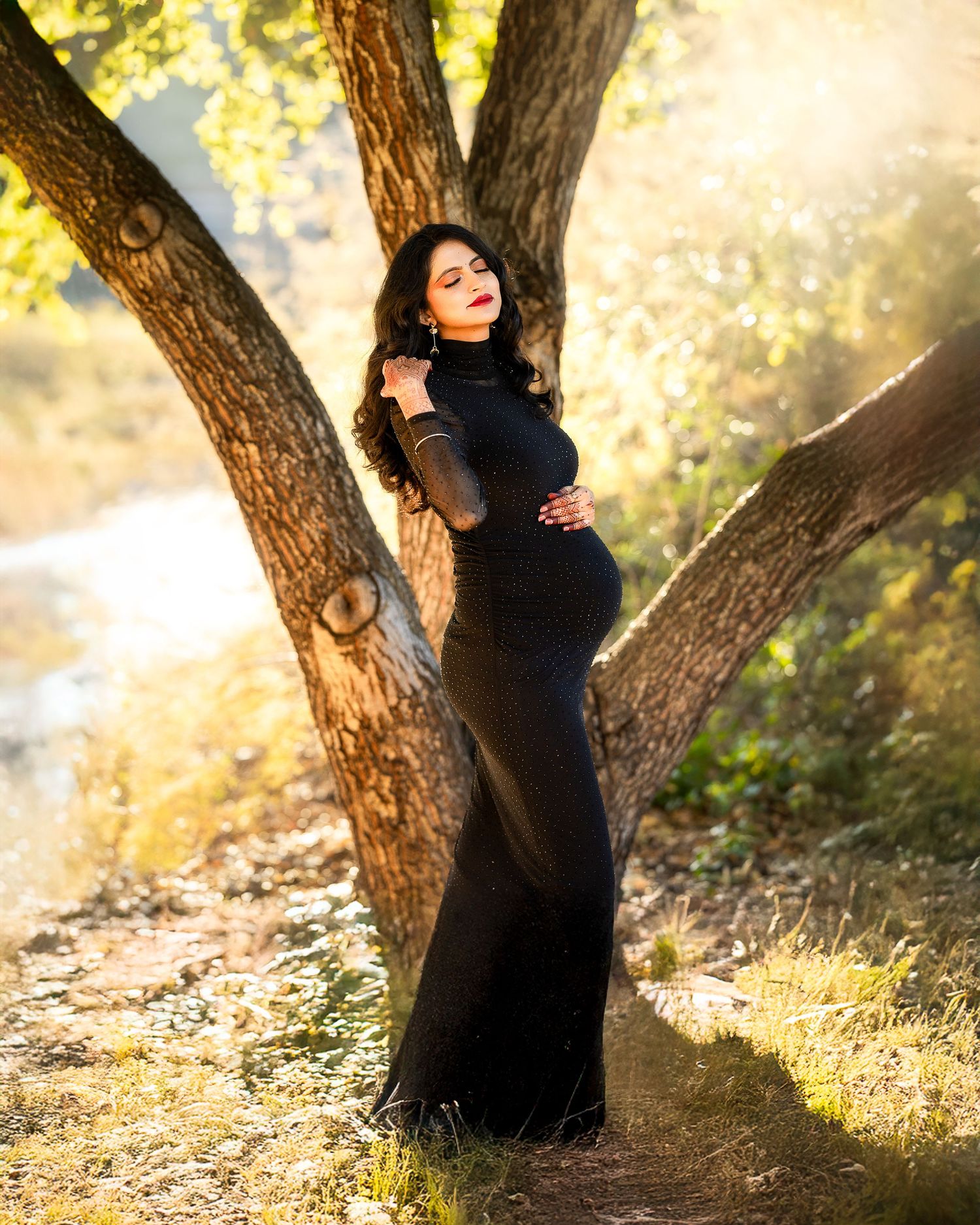 A maternity photoshoot in a dreamy outdoor setting with golden sunlight filtering through trees and tall grass.