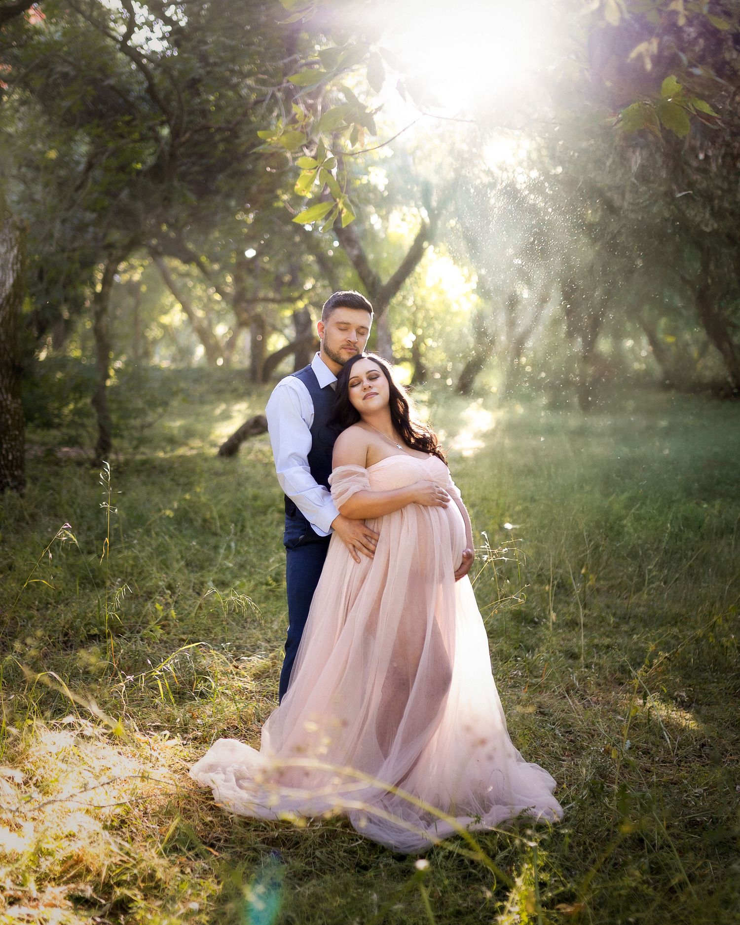 Couple poses in sunlit forest, pregnant woman in flowing pink gown embraced by man in navy suit.