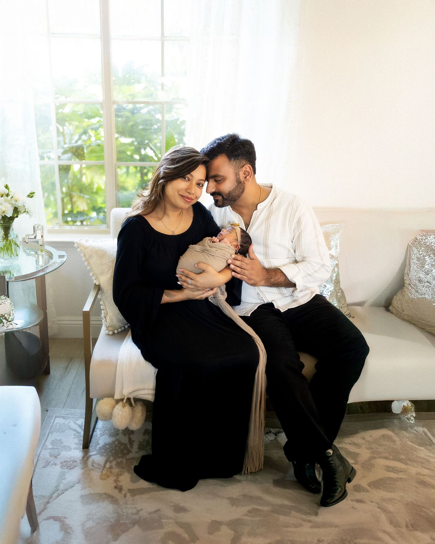 A couple in elegant attire tenderly gazes at their newborn baby while seated on a white sofa near a bright window.