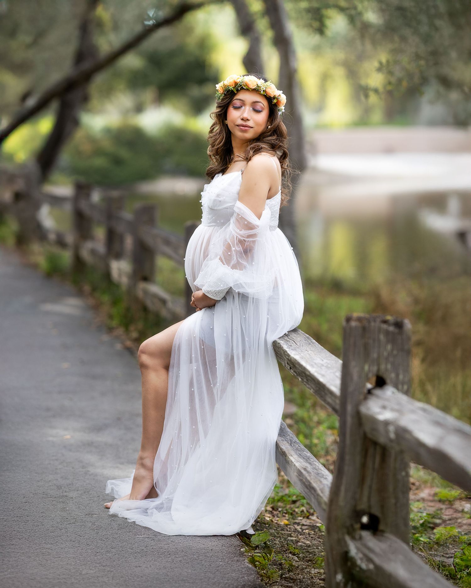 Ethereal maternity photo of a woman in flowing white dress and flower crown posing by wooden fence near water.