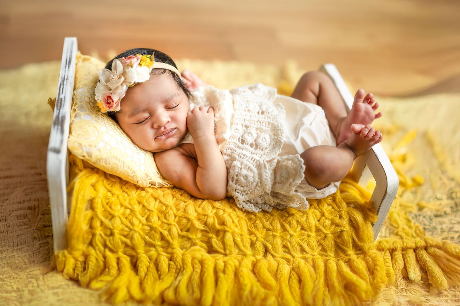 Newborn baby sleeping peacefully on yellow knit blanket in small white bed wearing lace outfit and floral headband.