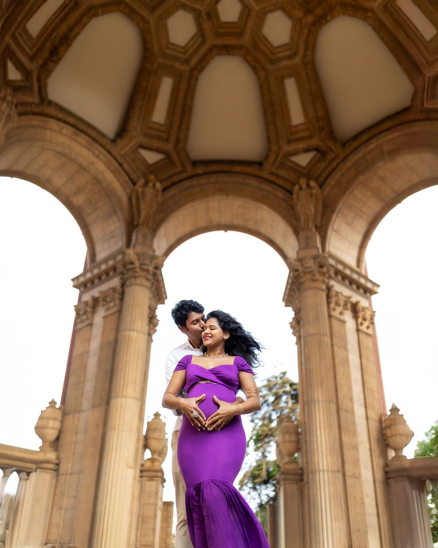 A person in a flowing purple gown poses elegantly beneath ornate stone arches with classical architectural details.