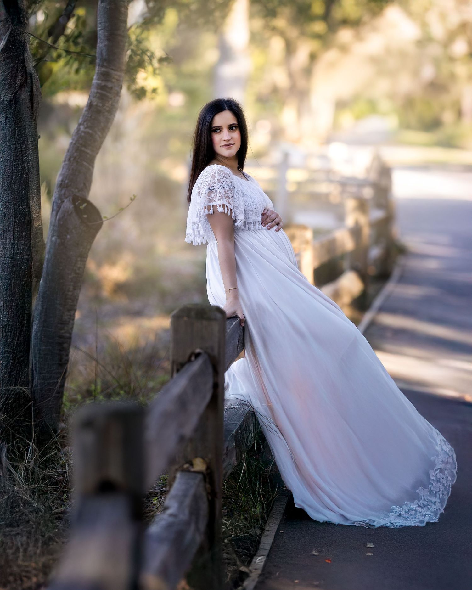 A flowing white lace maternity gown photographed outdoors against a sunlit path with natural bokeh effects.