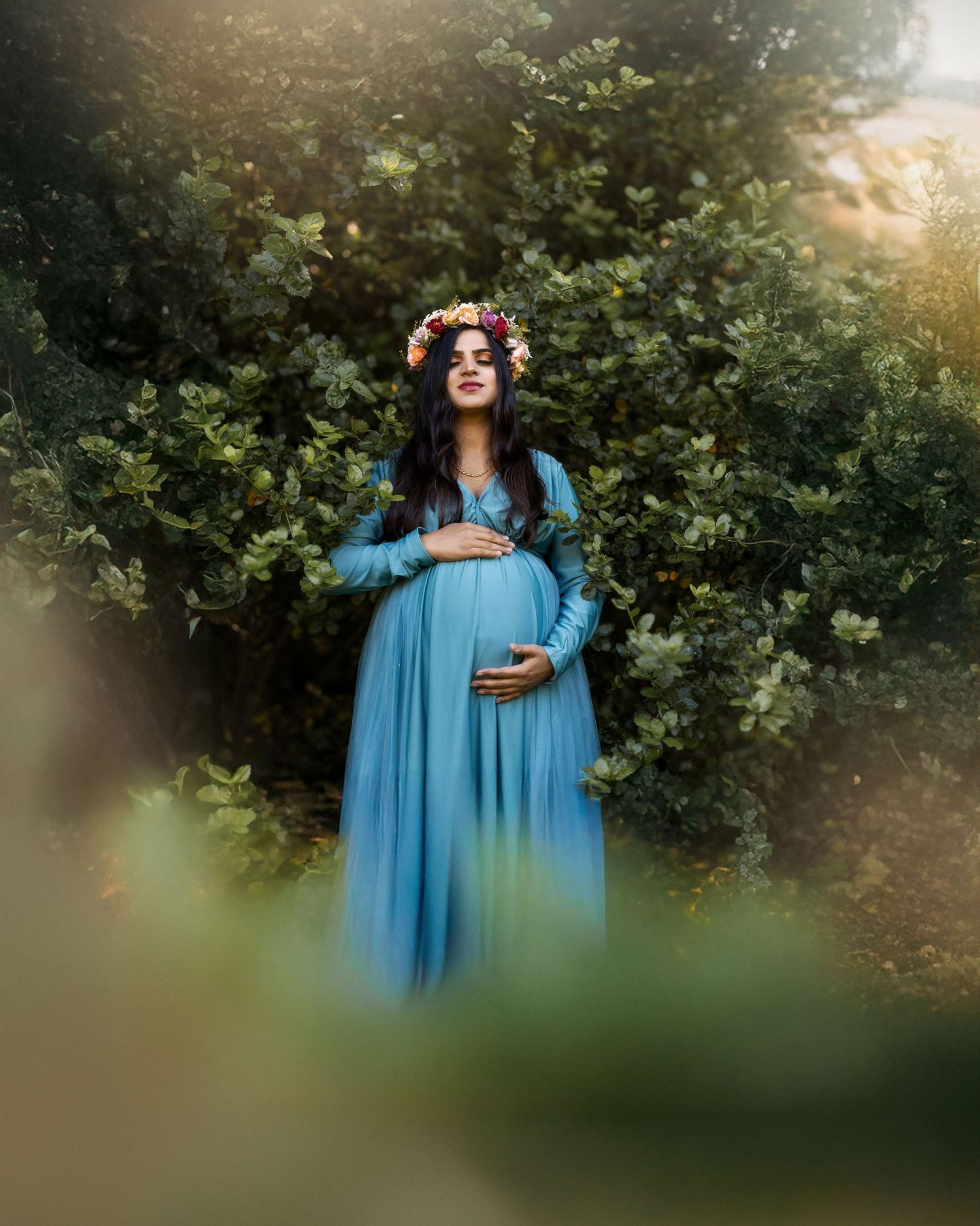 A person in a flowing light blue dress poses gracefully in a garden setting during golden hour.