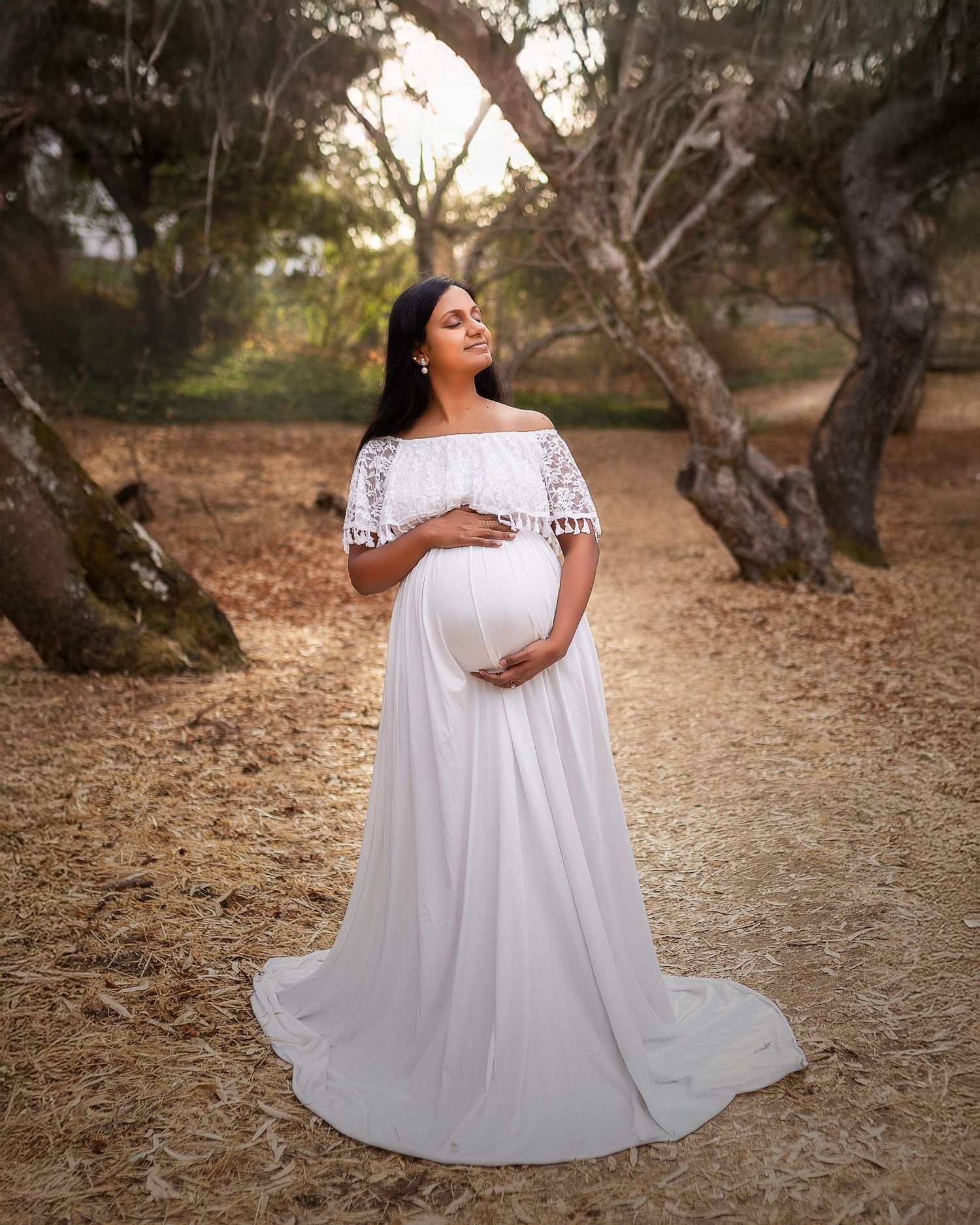 A person in a flowing white lace dress poses gracefully on a winding dirt path surrounded by rustic trees.