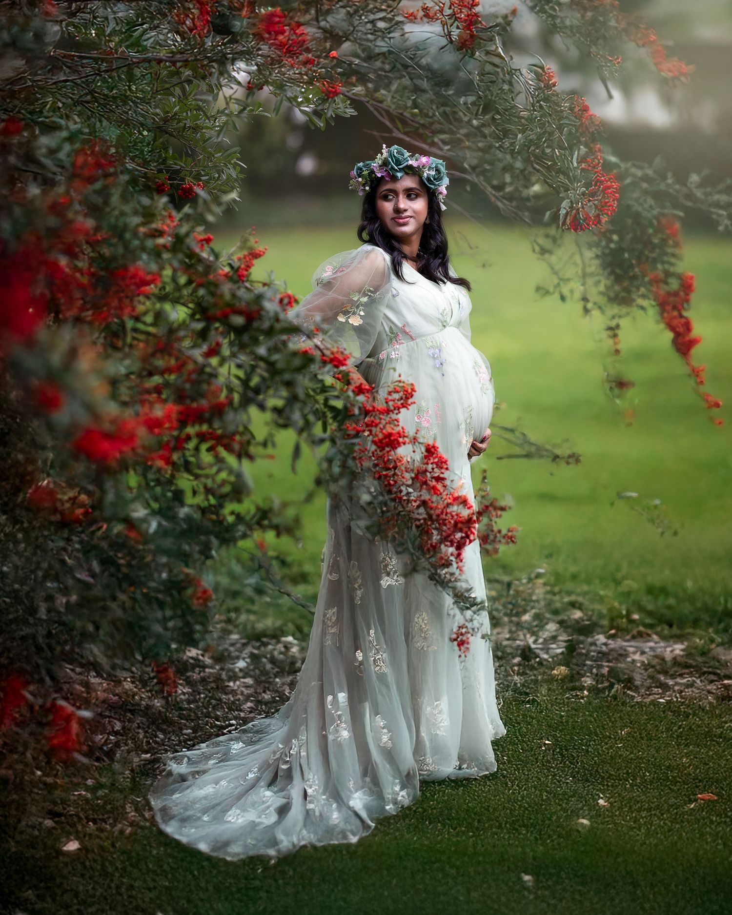 A pregnant woman in a flowing white lace dress and floral crown poses beneath red flowering branches in a dreamy garden setting.