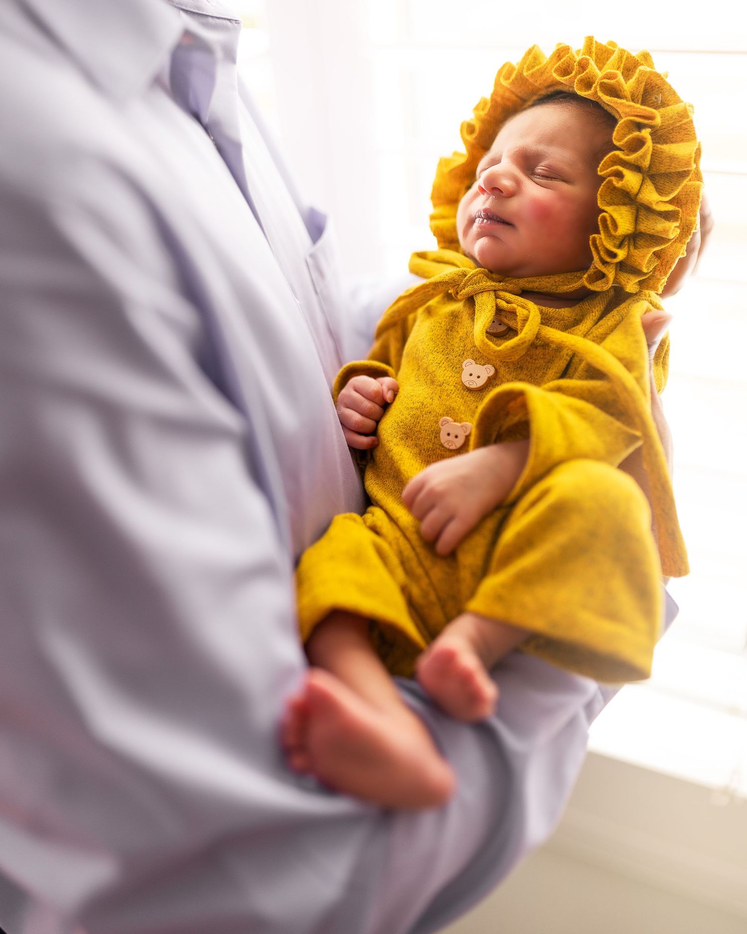 Sleeping newborn baby wearing a cozy yellow knit bonnet and sweater rests peacefully on soft gray blankets.