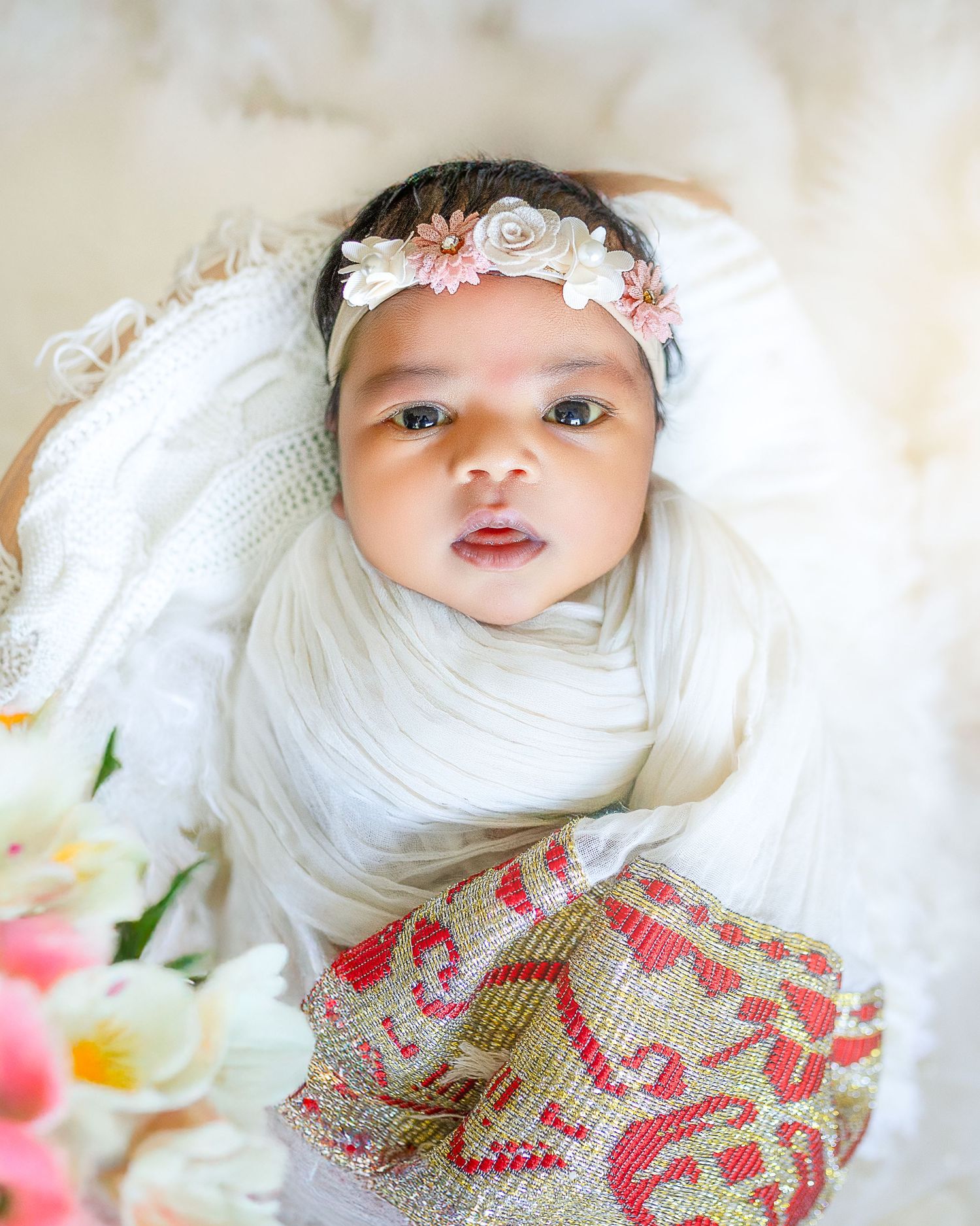 A baby wearing a white lace headband and floral dress lies on a light-colored blanket.