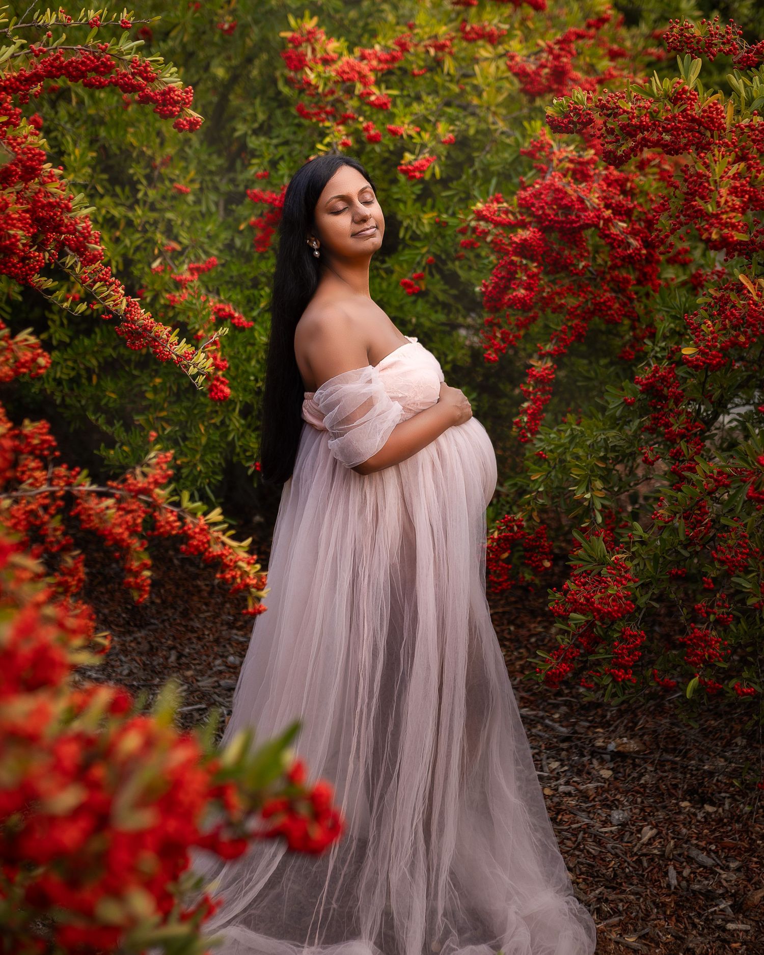 A dreamy maternity photo features a blush pink tulle gown surrounded by bright red autumn foliage in a garden.