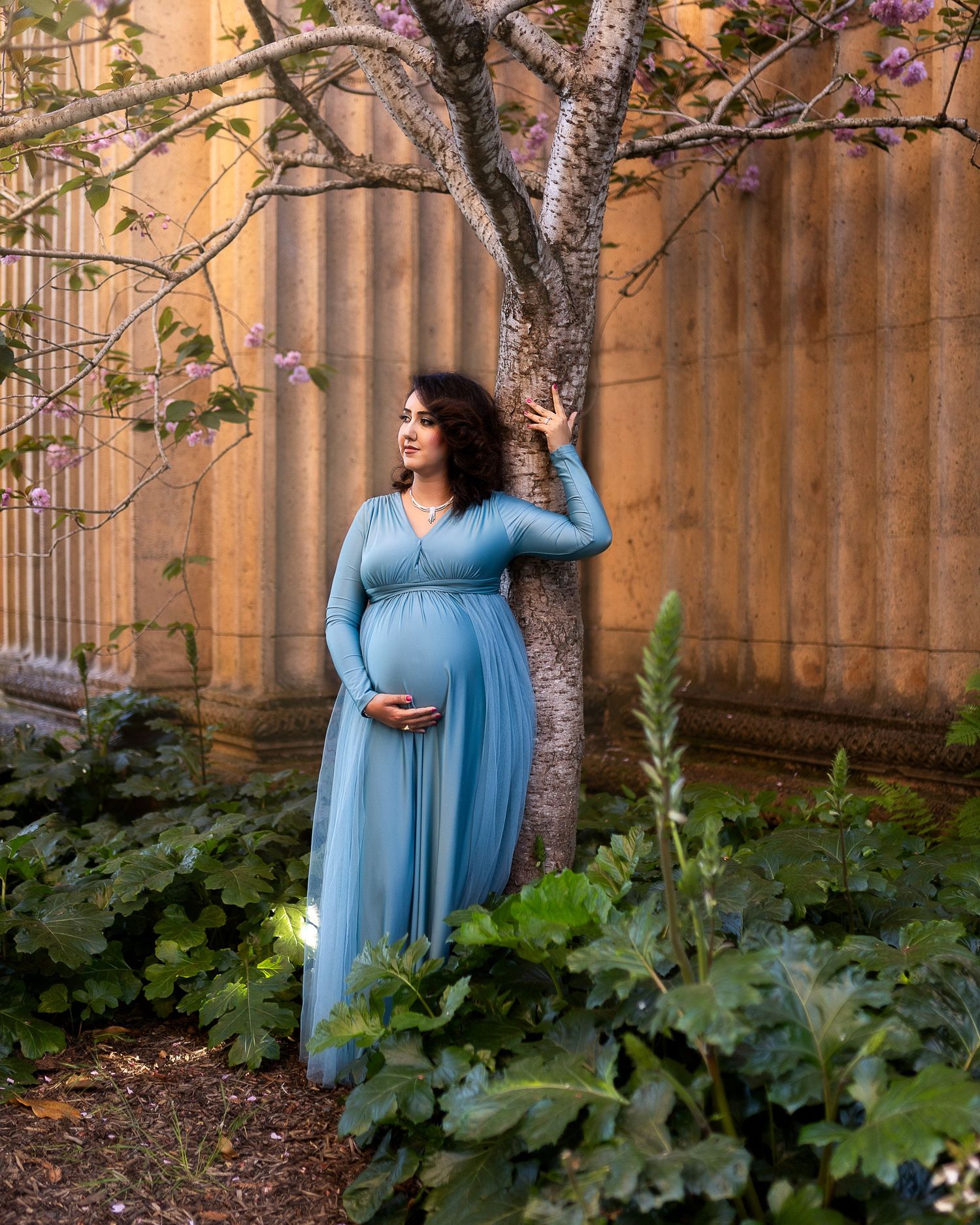 A woman in a flowing blue dress poses gracefully in a garden setting against a terracotta wall and lush greenery.