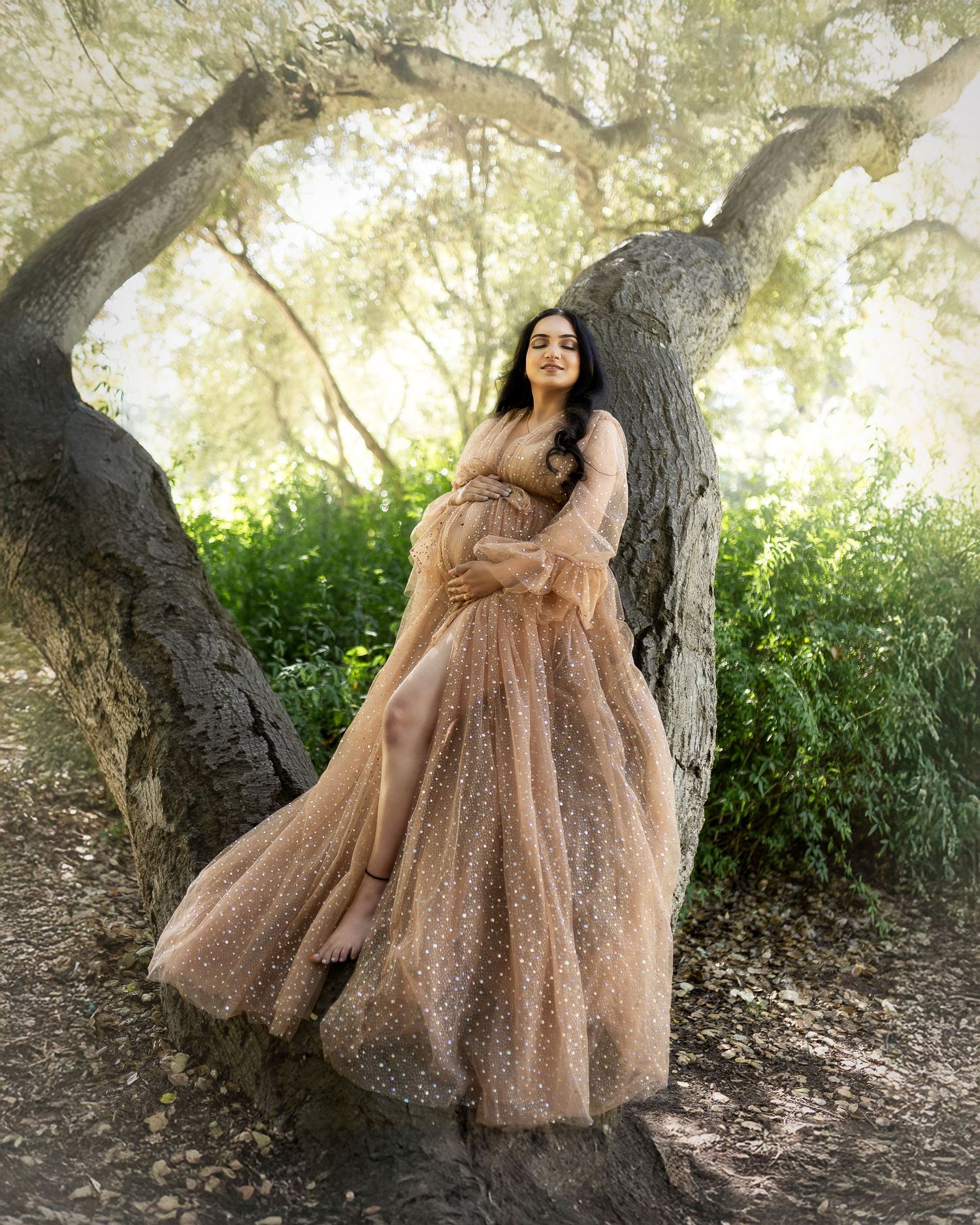 A person wearing a sparkly rose gold ballgown poses gracefully against a tree trunk in a natural outdoor setting.