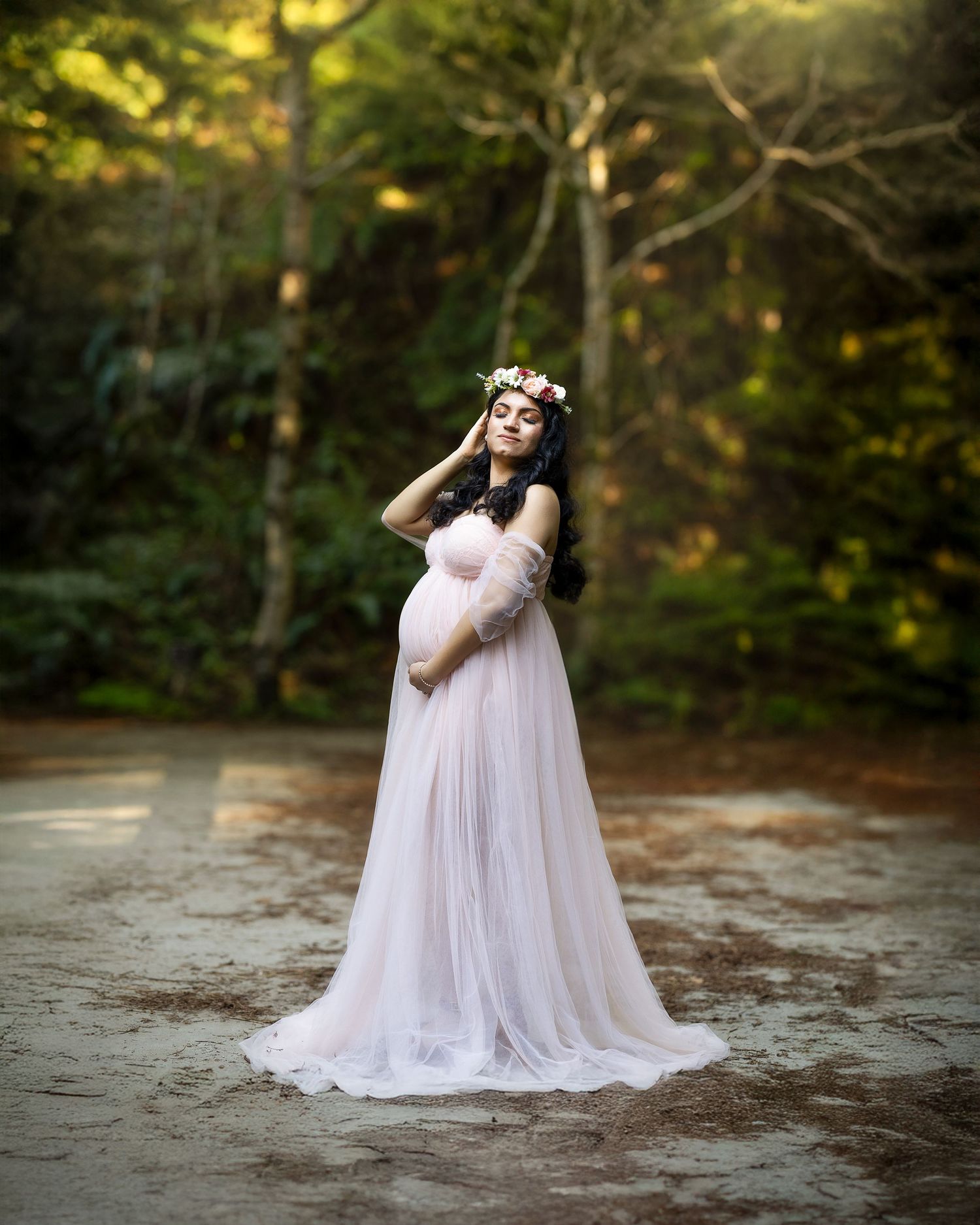 A figure in a flowing white lace maternity gown poses gracefully in a sunlit forest setting.