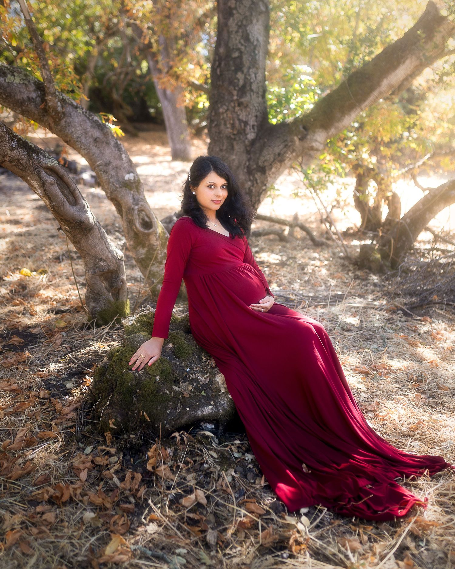 A person in a flowing burgundy dress poses gracefully among trees in dappled sunlight during an outdoor photoshoot.