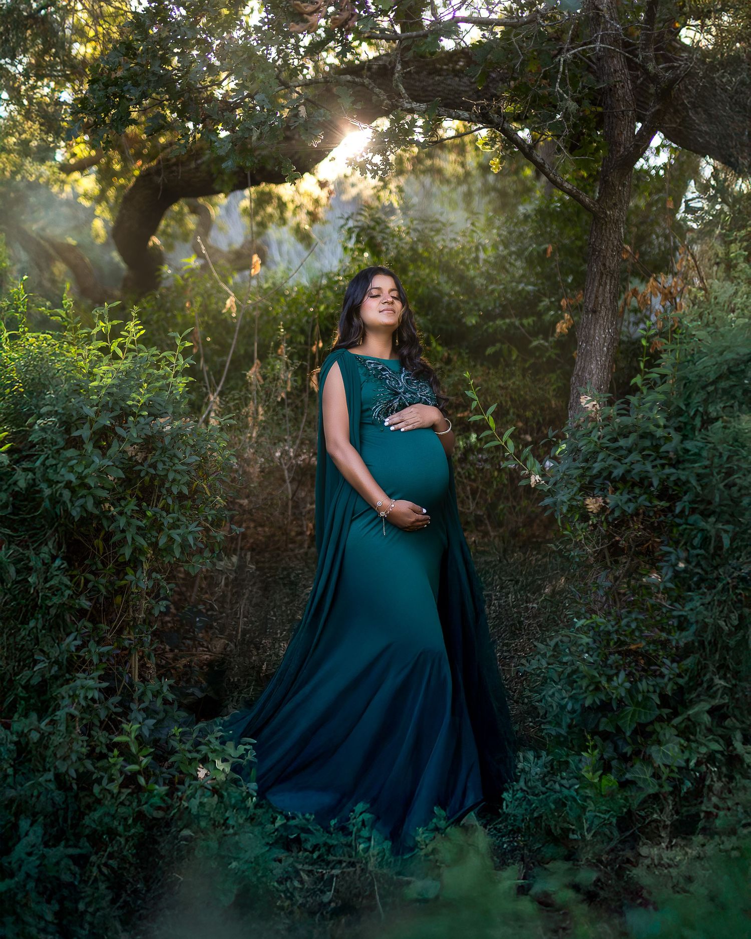 A flowing teal maternity dress catches golden sunlight in a dreamy outdoor forest setting.