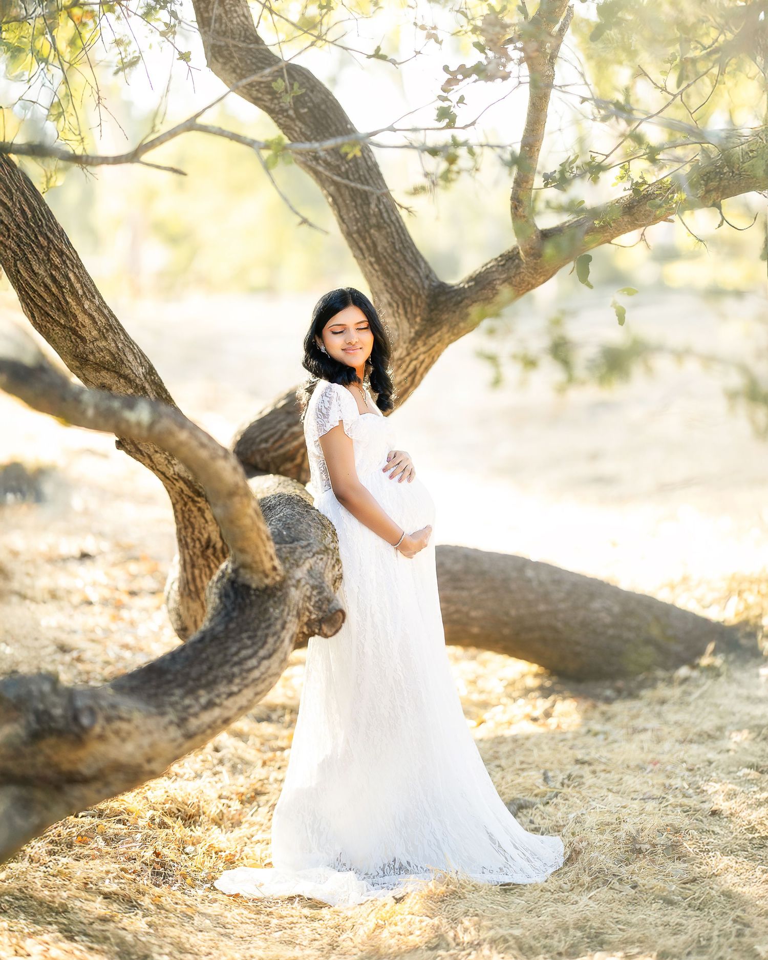 A person in a flowing white dress poses gracefully against a sunlit tree in a dreamy outdoor setting.