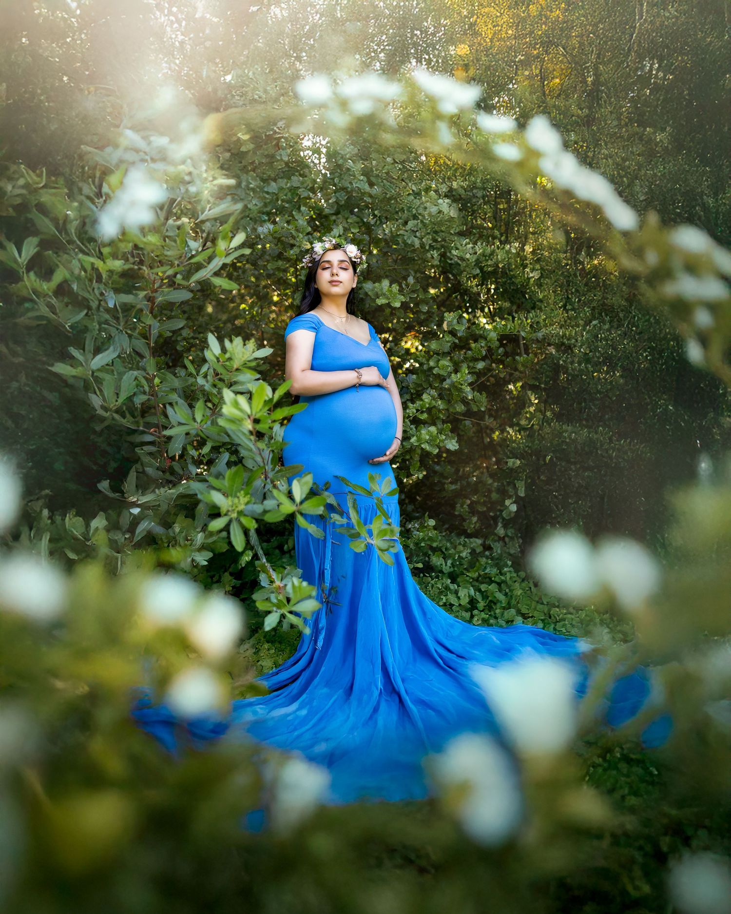 A flowing blue gown drapes elegantly in a dreamy garden setting surrounded by delicate white flowers and greenery.