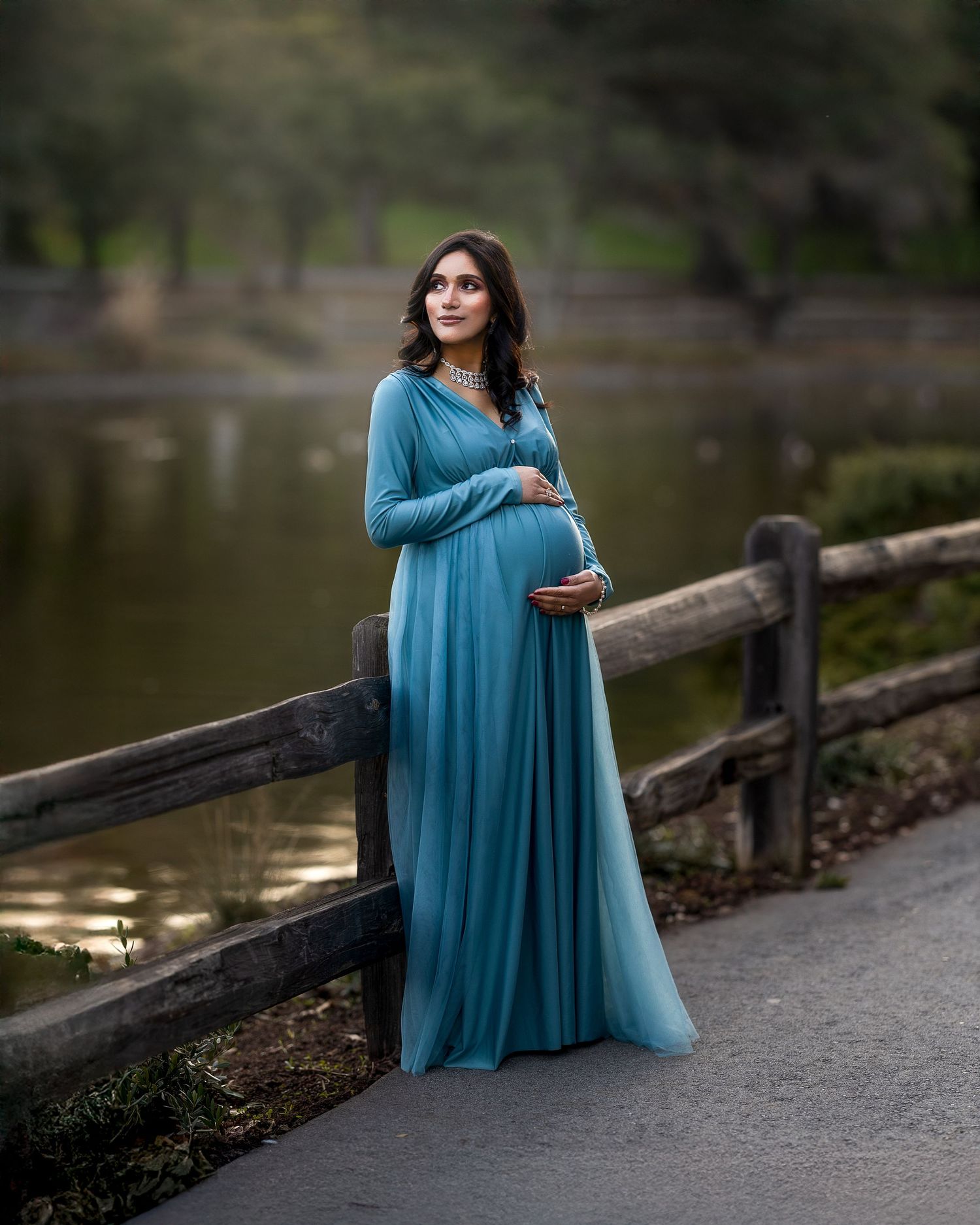 Peaceful maternity portrait by a lake with wooden railing and flowing blue dress at golden hour.