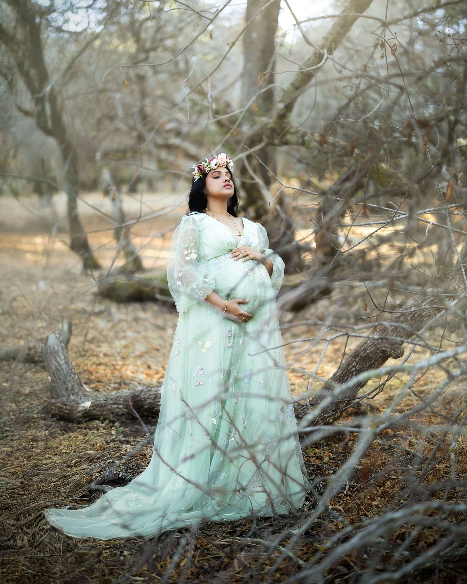 A dreamy maternity photo in a mint green dress among bare winter trees and branches in a forest setting.